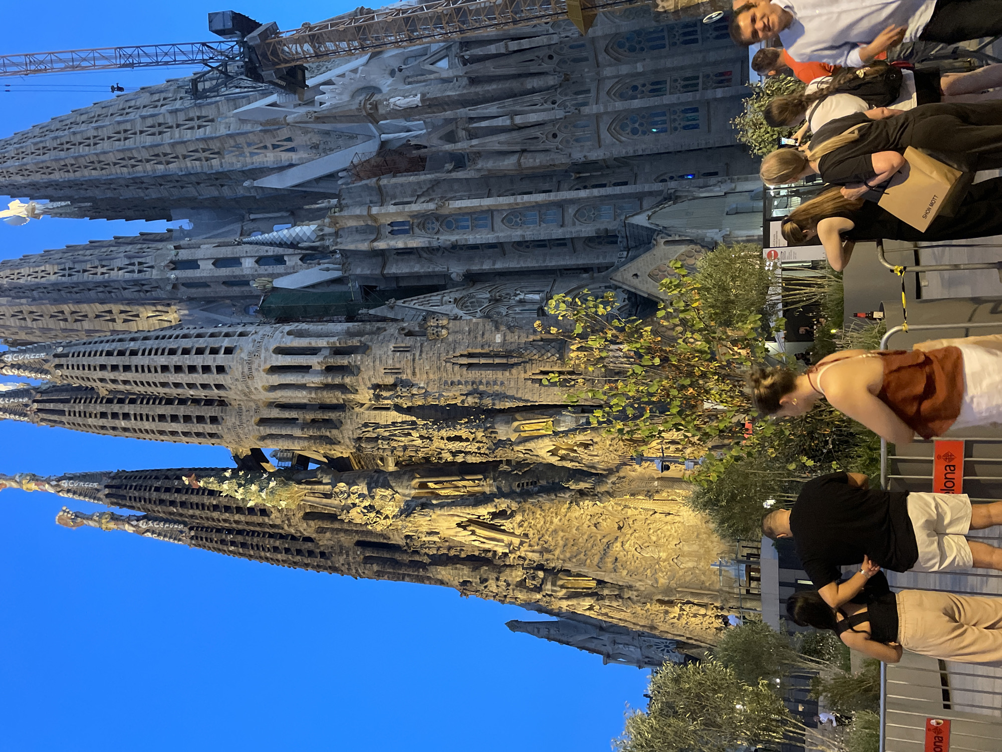 The image captures the Sagrada Familia basilica in Barcelona, Spain, at dusk. The basilica's iconic, intricate stone spires dominate the skyline against a deep blue sky. A construction crane is visible on the right side of the basilica, indicating ongoing work. In the foreground, a group of people are gathered, appearing to be tourists or onlookers. They are standing behind a metal barrier, with some individuals taking photographs. The lighting suggests the sun has set, with warm artificial lights illuminating the lower sections of the basilica. Trees and foliage are present in the middle ground, adding a touch of nature to the urban landscape. The overall scene is one of awe and admiration for the architectural marvel.