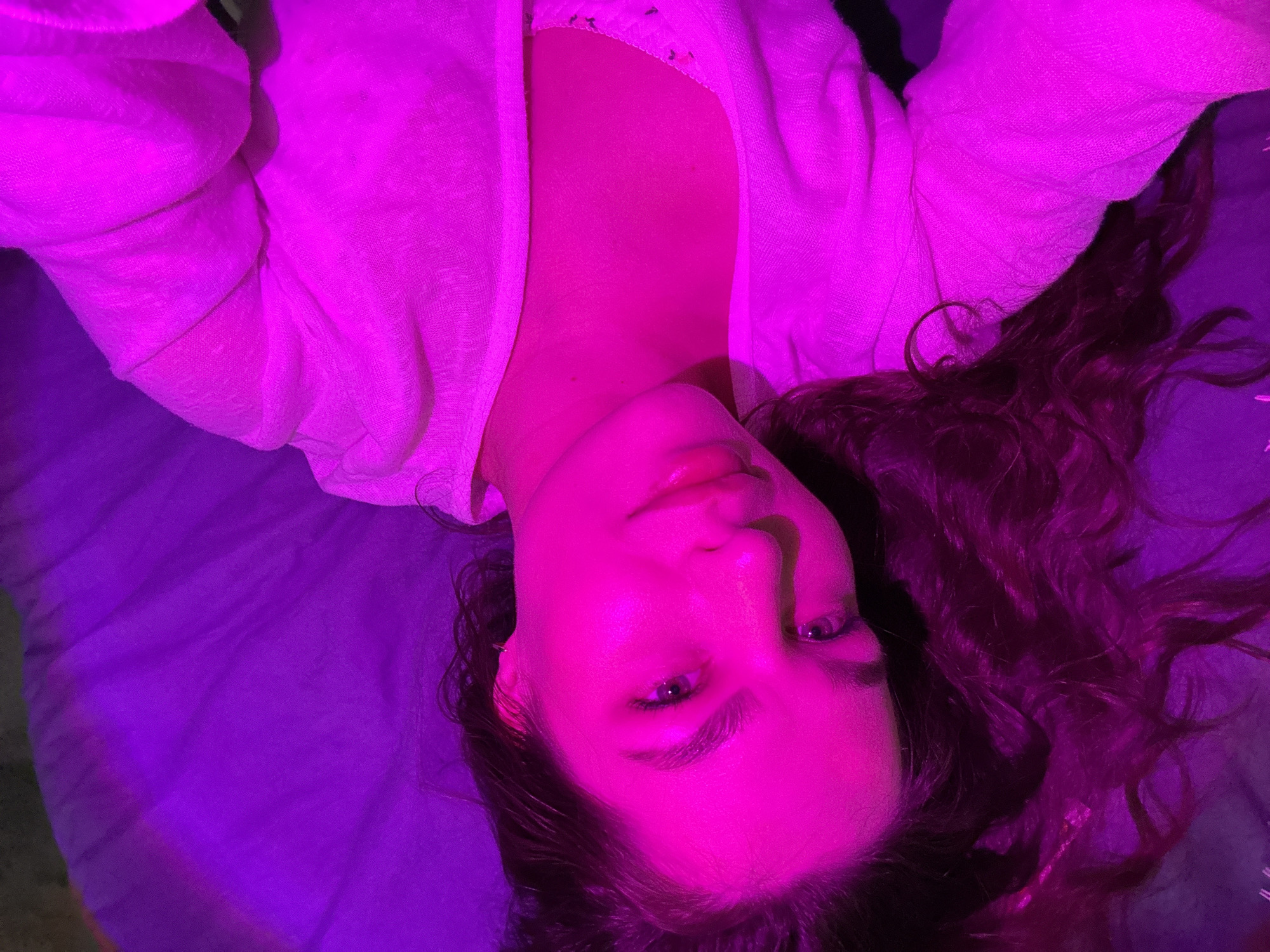 A woman lies on a bed in Chaikovsky, Russia. The lighting is predominantly pink and purple. The woman has fair skin, long curly brown hair, and is wearing a light-colored sweater or robe. She is looking directly at the camera with her eyes open. The background is a solid purple color, likely a bedsheet.
