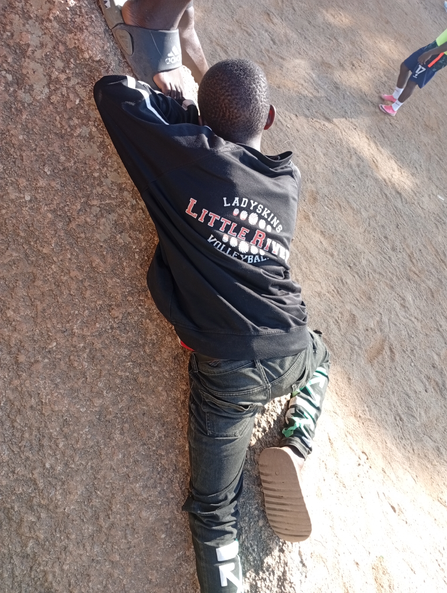 The image depicts a young boy wearing a black hoodie with "LADYSKINS LITTLE RIVER VOLLEYBALL" written on the back, dark jeans, and a patterned sock with green and white stripes. He is seen climbing a rocky surface, possibly a large boulder or hillside. His right foot is in a brown sandal. In the background, another person is partially visible, wearing blue shorts, pink sneakers, and a neon green top. The ground appears to be dry and sandy. The lighting suggests it is daytime, with shadows cast by sunlight. The rocky texture and the vegetation visible in the background are consistent with the natural landscape found in areas around Jos, Nigeria, known for its rocky outcrops and plateaus.