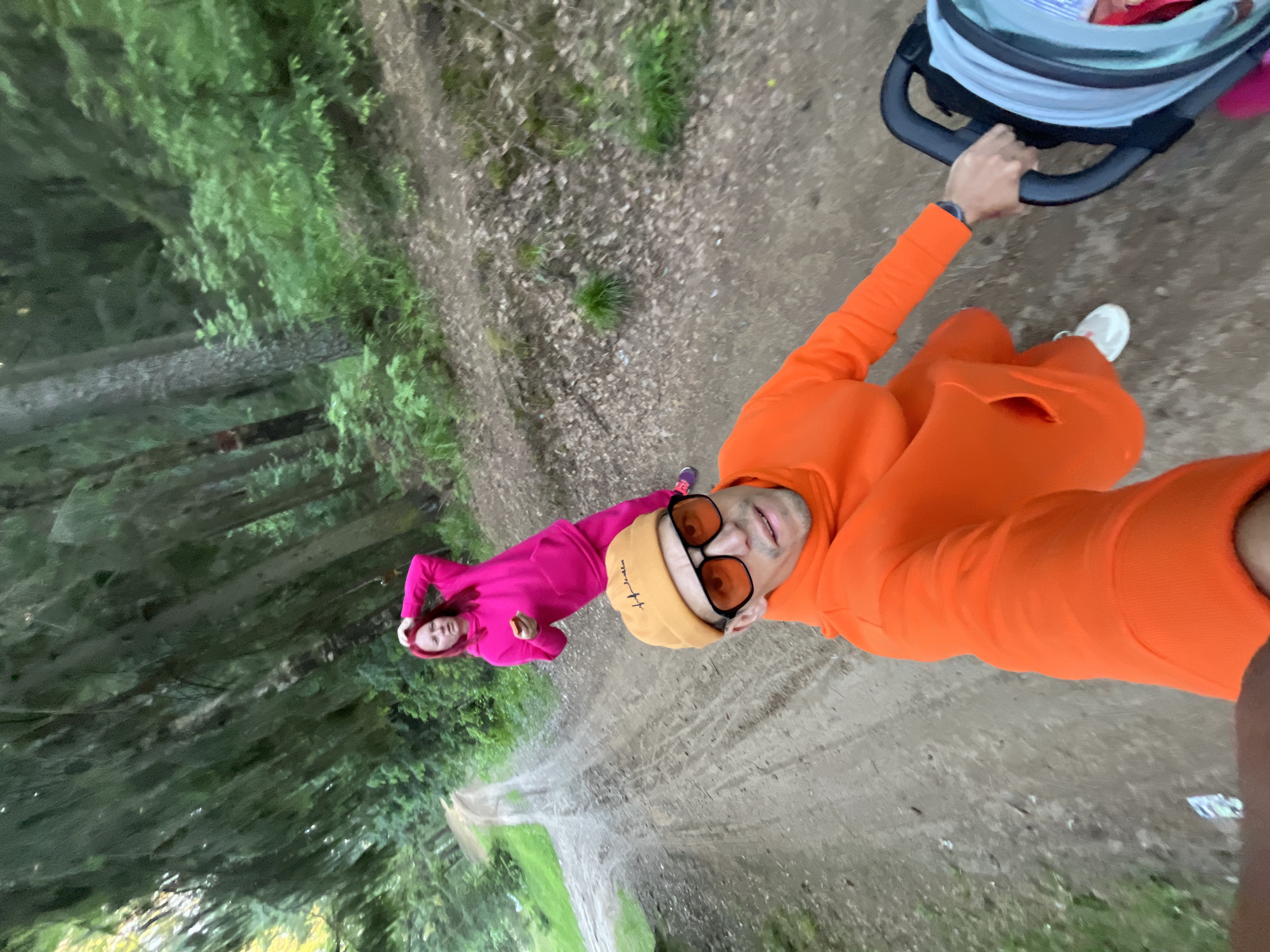 The image captures a selfie taken by a man pushing a baby stroller, with a woman walking behind him on a forest path, likely in Chaikovsky, Russia. The perspective is from the man's point of view, looking slightly upwards and towards the camera he is holding.
In the foreground, a man is visible from the chest up, wearing a vibrant orange tracksuit, an orange beanie, and orange-tinted sunglasses. White text, "Love for Freedom," is inscribed on his beanie. He has a watch on his left wrist and is holding the black handle of a light blue and grey baby stroller with his right hand. His expression is neutral to slightly smiling. His left leg and white sneaker are partially visible on the right side of the frame.
Further down the dirt path, a woman is seen walking. She is dressed in a bright pink tracksuit and has reddish hair, looking towards the camera.
The setting is a winding dirt or gravel path flanked by tall, dense evergreen trees, suggesting a forest or wooded area. Patches of green grass and scattered dry leaves are visible on the ground beside the path. The lighting appears soft and diffused, indicating a cloudy day or the shade of the forest. The overall scene conveys a leisurely stroll in a natural, serene environment.