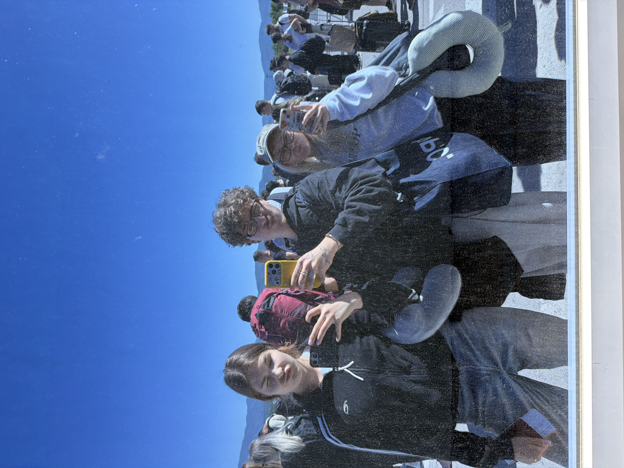 The image shows a group of people taking a selfie outdoors on a sunny day, likely at an airport in Gornje Dubrave, Bosnia and Herzegovina. Three individuals are the primary focus: a woman in a black jacket holding a phone, a man with glasses also holding a phone, and a woman in a light blue sweatshirt and hat. Other people, some with luggage, are visible in the background. The sky is clear and blue. The photo appears to be a reflection in a glass surface.