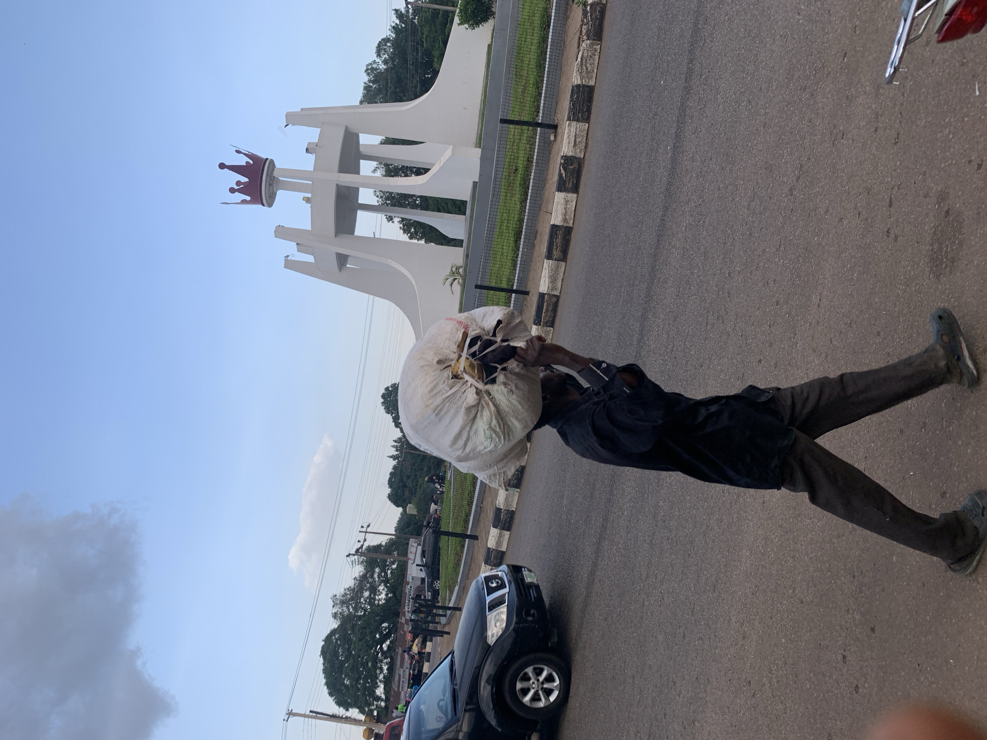 A person walks down a paved street in Jos, Nigeria, carrying a large sack of goods on their head. The person is wearing dark clothing and dark shoes. In the background, a white monument with a red crown sits atop a circular island. Cars are present on the street, and trees and power lines are visible in the distance. The sky is partially cloudy.