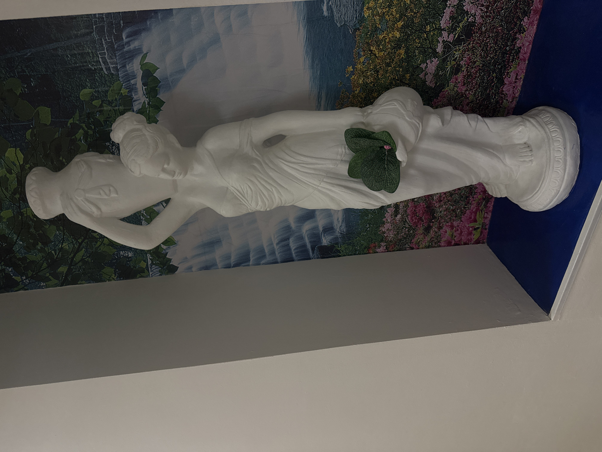 A white statue of a reclining nude woman is displayed against a background of a waterfall and flowers. The statue is lying on its side on a blue platform. The statue is holding a bunch of green leaves with a small pink flower in her left hand. The background is a print of a waterfall flowing through a forest, with some areas showing flowering plants. The statue is positioned in a niche with a white trim and is set against a white wall. The location is not specified, and there is no information that places the scene in Yevpatoriya, Ukraine.