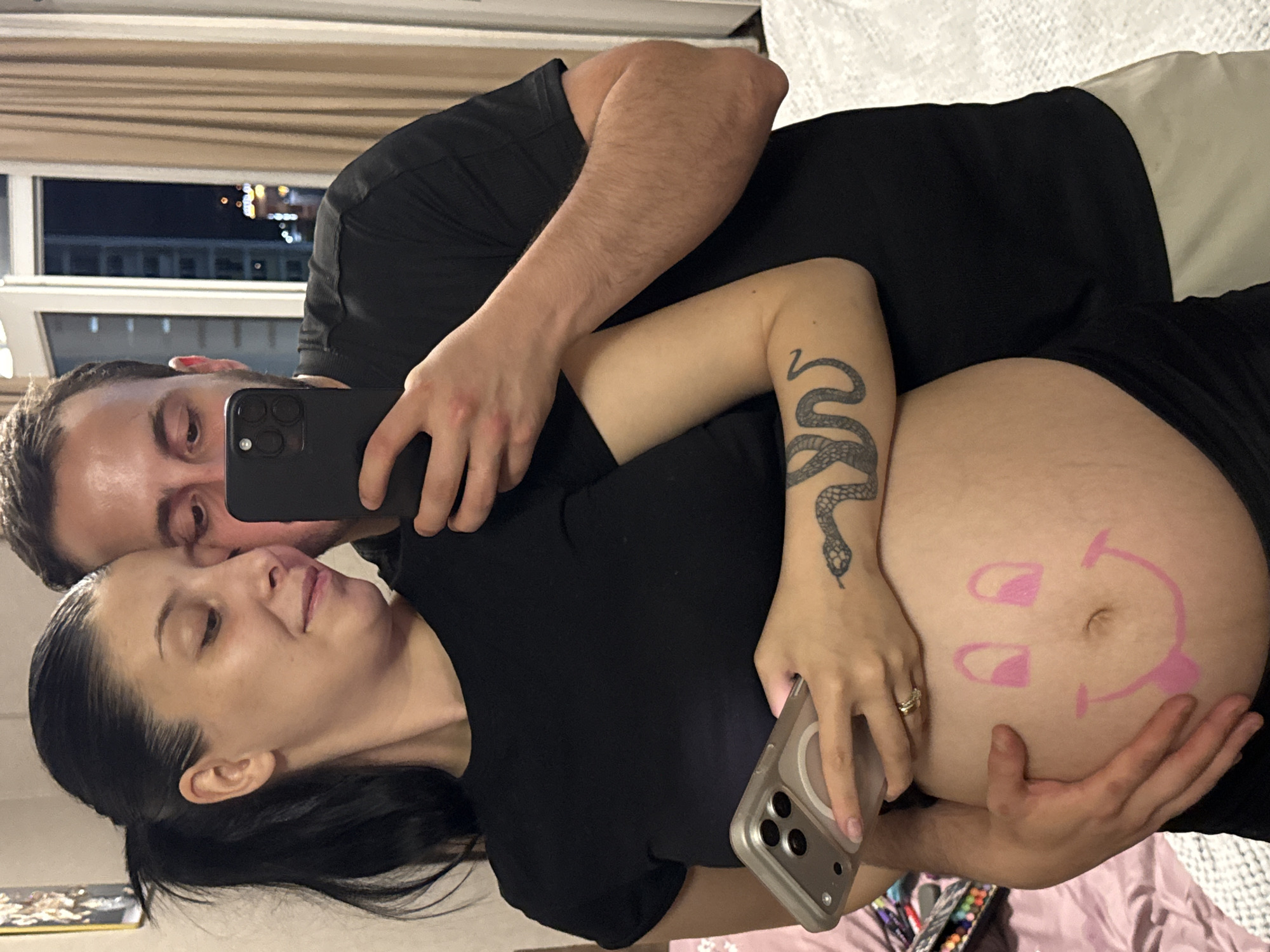 A pregnant woman and a man are posing for a selfie. The woman is wearing a black t-shirt, and the man is wearing a black t-shirt. The woman's belly is painted with a smiley face. The man is holding a phone to take the photo. They are standing in front of a window with a view of a city at night, which is likely Tbilisi, Georgia, given the architectural style of the buildings and the general lighting conditions. The room appears to be a bedroom, with a bed and curtains visible. The lighting is dim, with the primary light source coming from the city lights outside and the phone's screen.