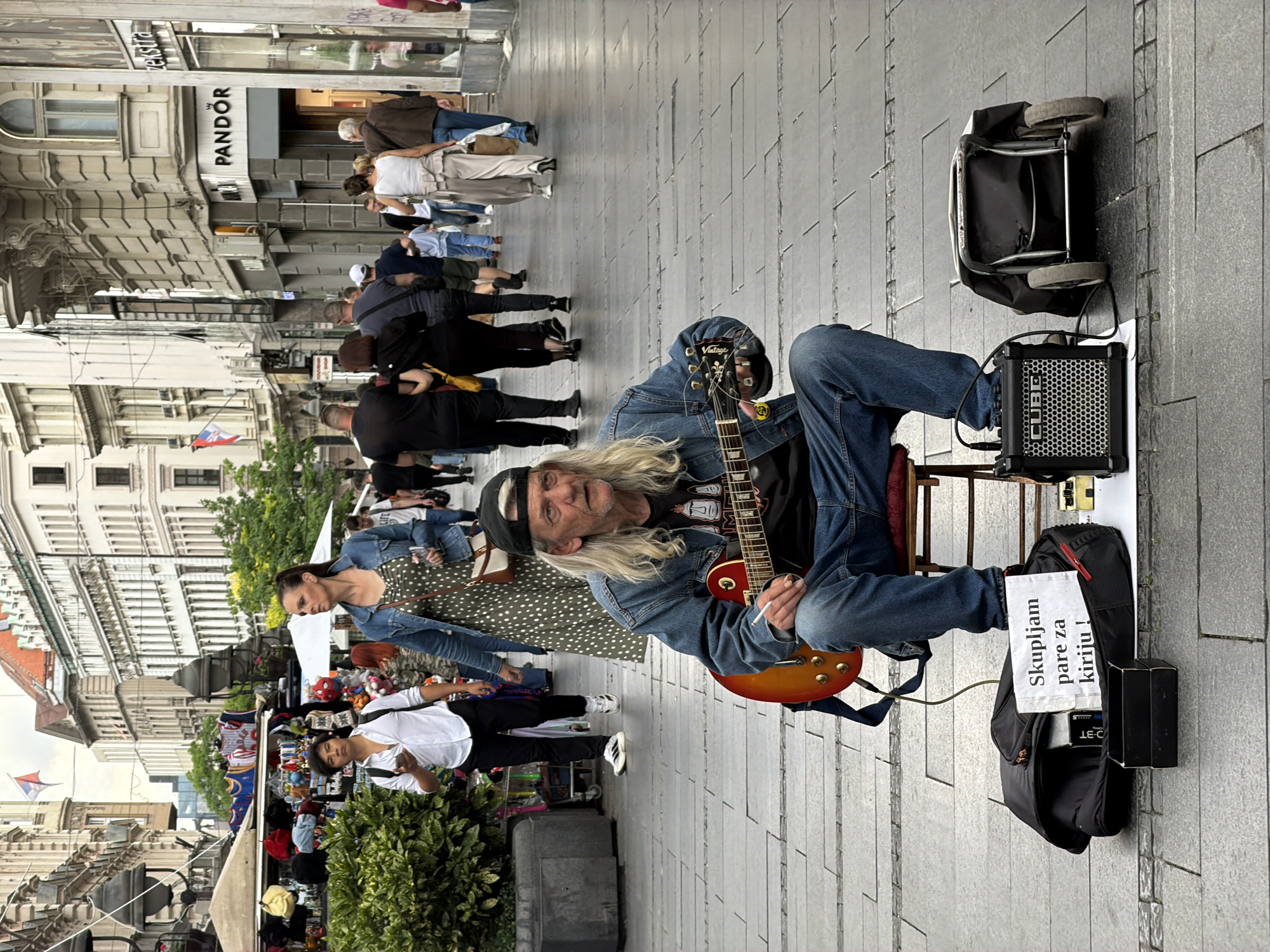 The image captures a street scene in Belgrade, Serbia. A street musician is seated on a small stool, playing an electric guitar. He wears a denim jacket, jeans, and a black cap. His long gray hair is visible. A small amplifier and a trolley are positioned beside him. A handwritten sign in Serbian reads, "Skupljam pare za kiriju!" ("I'm collecting money for rent!"). A crowd of people walks by, some glancing at the musician. Buildings line the street, including one with a Pandora store. A vendor selling various items can be seen in the background. The street is paved with light gray stones.