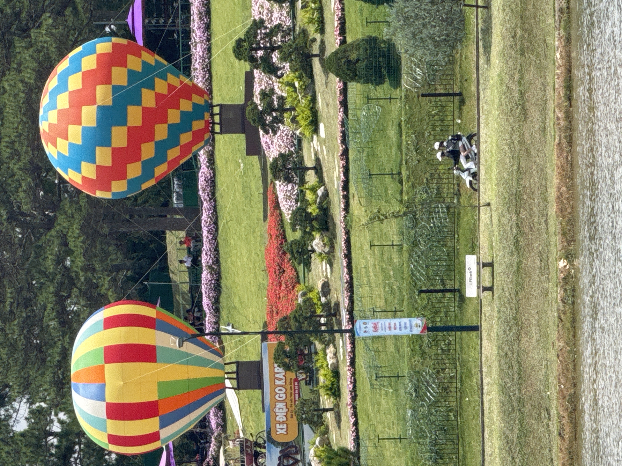 The image captures a scenic view in Da Lat, Vietnam, likely at the Quang Trung Flower Park or a similar botanical garden. The foreground features a tranquil body of water, with a grassy embankment leading up to a manicured landscape. Two large, colorful hot air balloons, one with vertical stripes and the other with a checkered pattern, are prominently displayed as attractions. The balloons are tethered and positioned on platforms.
The midground is a lush, tiered garden filled with vibrant red and pink flowers, interspersed with meticulously shaped bonsai trees and ornamental shrubs. A pathway meanders through the garden. In the background, tall pine trees dominate the skyline, characteristic of Da Lat's mountainous terrain.
On a paved path in front of the garden, two individuals are riding a scooter, wearing helmets, suggesting a mode of transport or a leisurely ride within the park. To the left, a sign with Vietnamese text, "XE ĐIỆN GO KART" (Electric Go-Kart), indicates an activity offered. Further to the left, a decorative cart is visible. A white bench with the "LPBank" logo sits on the grass, offering a place to rest. A tall, slim signpost with further Vietnamese text stands near the center of the image. The overall atmosphere is bright and sunny, with a sense of recreational activity and natural beauty.