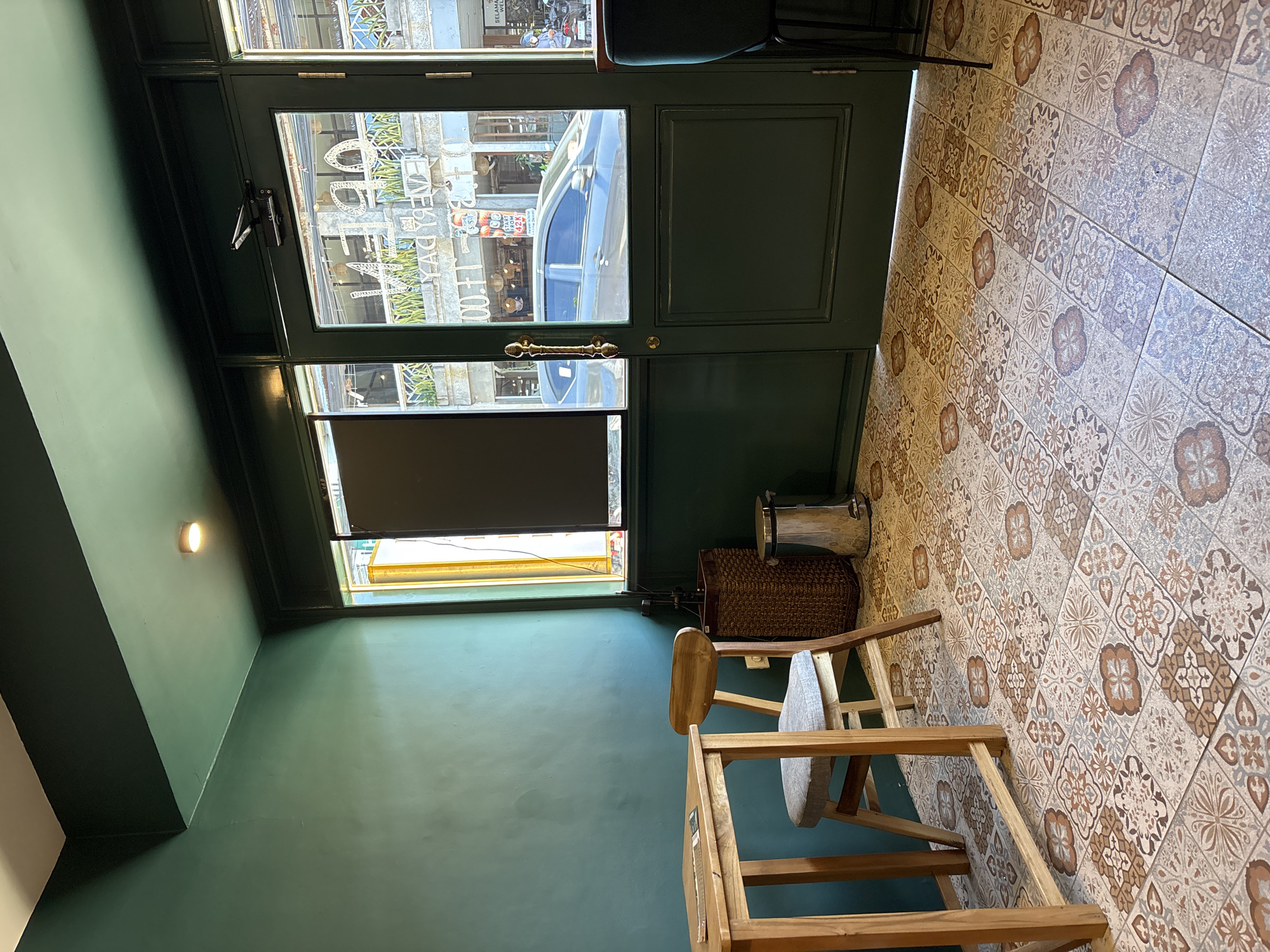 The image shows the interior of a shop or cafe in Kuta Selatan, Indonesia. The view is from inside, looking towards the entrance. The entrance consists of a glass door and a solid green door, both framed in dark green. The glass door has a metallic handle. There's an awning or blind partially drawn above the door. Outside the glass door, the street scene is visible, with buildings, a vehicle, and signs in Indonesian script.
To the right, the floor is covered with patterned tiles in earthy tones. A wooden chair with a gray cushion sits near a basket and a metallic trash bin. The left side of the image shows a wall painted in dark green, with a single, round light fixture.