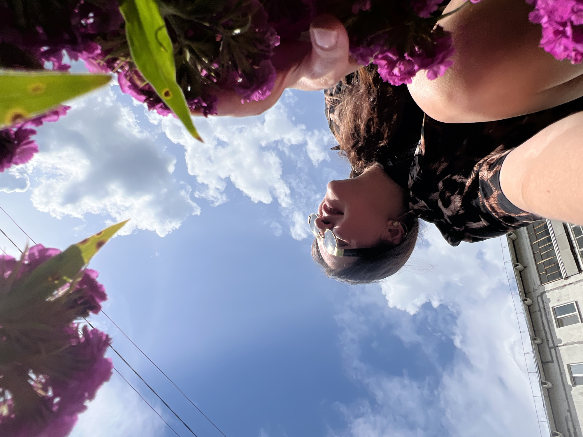 This is a low-angle, upside-down, close-up photograph taken outdoors. The primary subject is a person, likely female, viewed from below and looking upwards towards the sky. Her head is turned to the side, revealing her profile with sunglasses on. She has dark, curly hair and is wearing a patterned top. A hand is visible, reaching towards the foreground.
The setting appears to be a partly cloudy day with a bright blue sky. In the foreground and framing the shot are vibrant purple flowers and green leaves, suggesting a garden or an area with blooming plants. In the background, a concrete building with multiple windows and balconies is visible. Power lines are also present, stretching across the sky. The lighting indicates it is daytime, likely mid-morning or afternoon, given the brightness of the sky. The overall mood is relaxed and possibly contemplative, with the subject looking up into the expanse of the sky, surrounded by nature and urban elements. The image is taken in Noviy, Russia. No visible text is present in the image.
