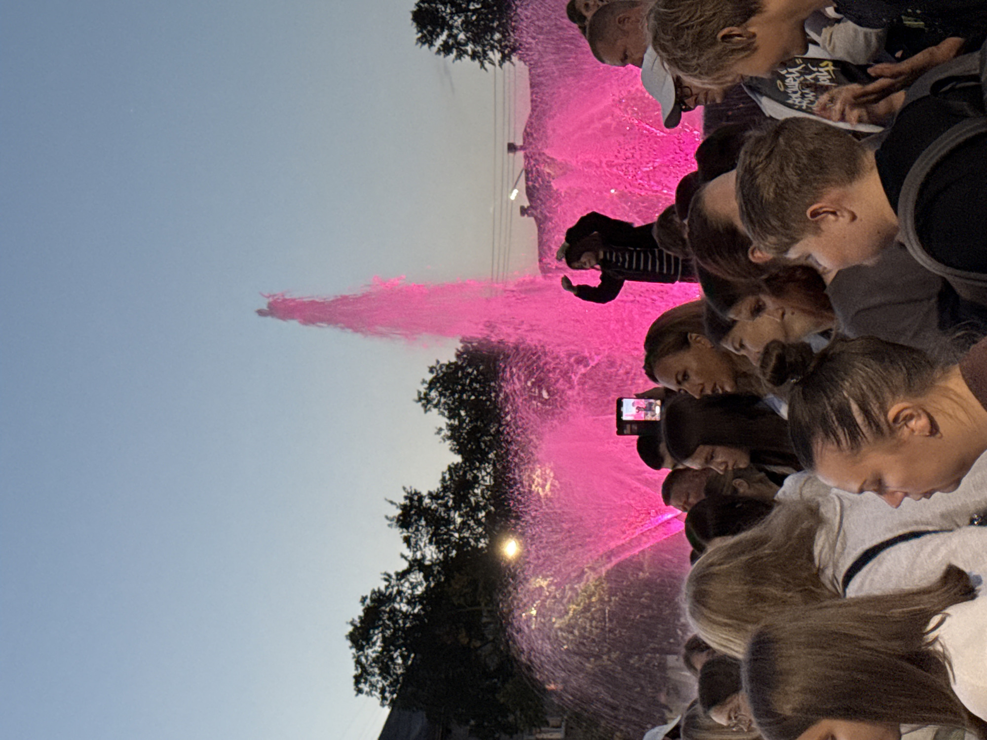 In Chaikovsky, Russia, a crowd of people is gathered in front of a brightly lit pink fountain during the twilight hours. The fountain sprays a tall, vibrant stream of water upwards, illuminated by pink lights. Among the spectators, a young person in the foreground is captured on a mobile phone, possibly taking a photo or video of the spectacle. The sky above is a gradient of pale blue and orange, indicating the fading light of day. The scene suggests a public event or gathering, likely a celebration or a popular attraction in the city.