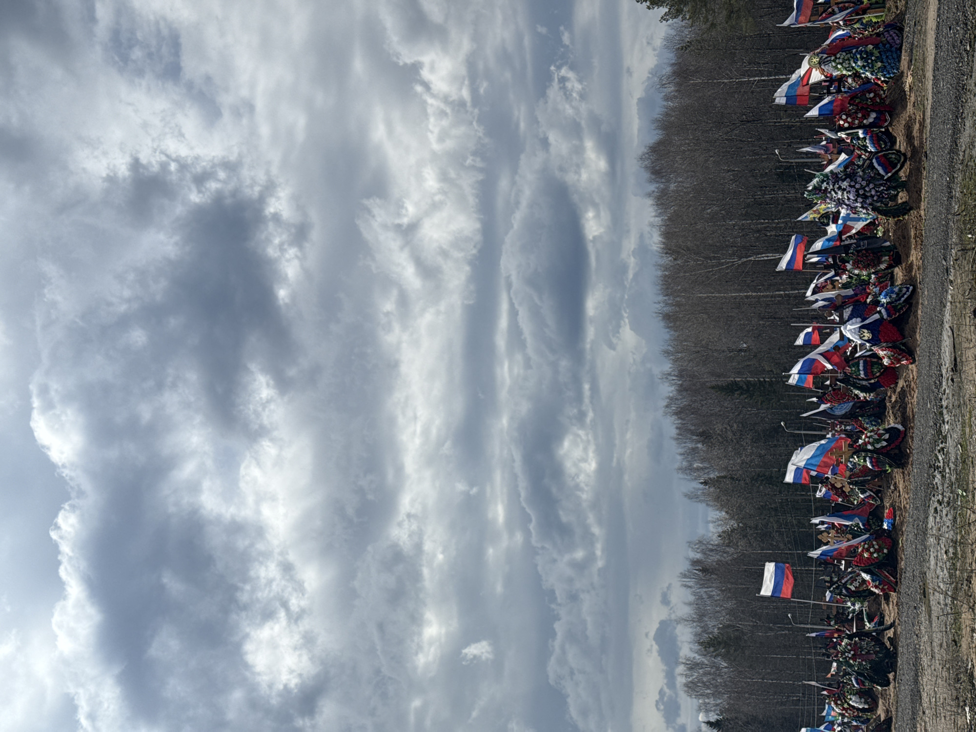 The image, taken in Perm, Russia, depicts a solemn outdoor scene, likely a military cemetery or a memorial site. A long line of graves or memorials stretches across the middle ground, bordered by a dirt path in the foreground and a dense forest of bare, leafless trees in the background. The sky, dominating the upper portion of the image, is heavily overcast with dramatic, thick grey and white clouds, suggesting a somber atmosphere and a cold season such as late autumn or early spring.
Each memorial is adorned with a variety of colorful floral wreaths, some featuring Orthodox Christian crosses. Prominently displayed at almost every memorial are numerous Russian flags, recognizable by their white, blue, and red tricolor design. Some flags also appear to bear additional military or national symbols, reinforcing the patriotic and commemorative nature of the site.
No people are visible in the photograph, implying a quiet moment of remembrance. The uniform arrangement of the memorials and flags suggests a dedicated, maintained memorial park for fallen soldiers or national figures. The bare trees in the background further contribute to the subdued and reflective mood of the scene. The lighting suggests it is daytime, but the heavy cloud cover prevents direct sunlight. No discernible text is visible on any of the objects in the image. The overall impression is one of respect, commemoration, and national reverence.