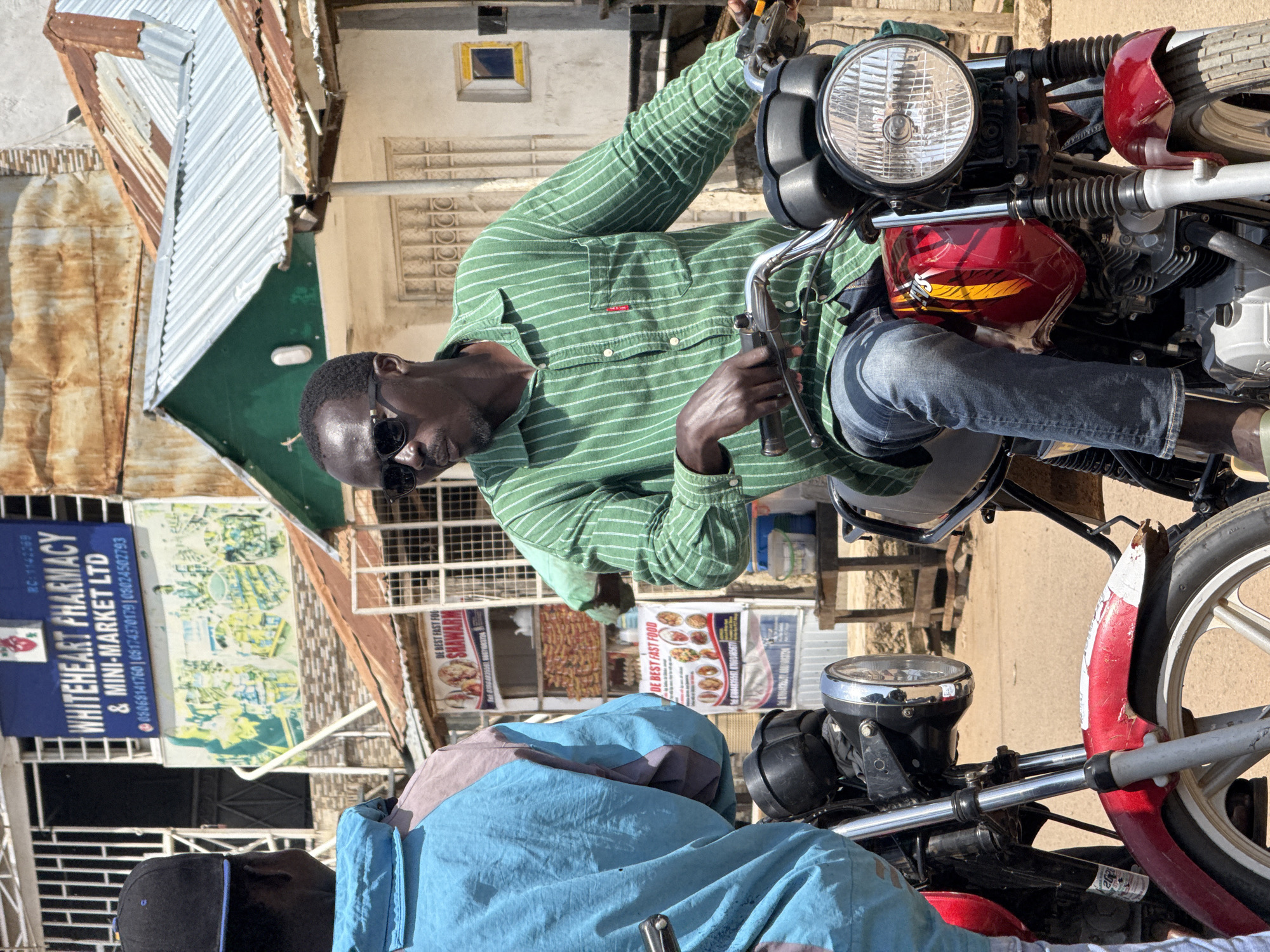 Two men are standing next to their motorcycles in a marketplace setting in Abuja, Nigeria. The man on the right is wearing a green and white striped long-sleeved shirt, blue jeans, and sunglasses. He is sitting on his red and yellow motorcycle, holding the handlebars. The man on the left is wearing a light blue and pink jacket and a black baseball cap. He is standing next to his red motorcycle, facing away from the camera. In the background, there are buildings with signs that read "WHITEHEART PHARMACY & MINI-MARKET LTD" and "DE BEST FAST FOOD SHAWAMA." There are also food stalls and other market activity visible. The ground is dusty, suggesting a dry climate. The lighting indicates it is daytime.