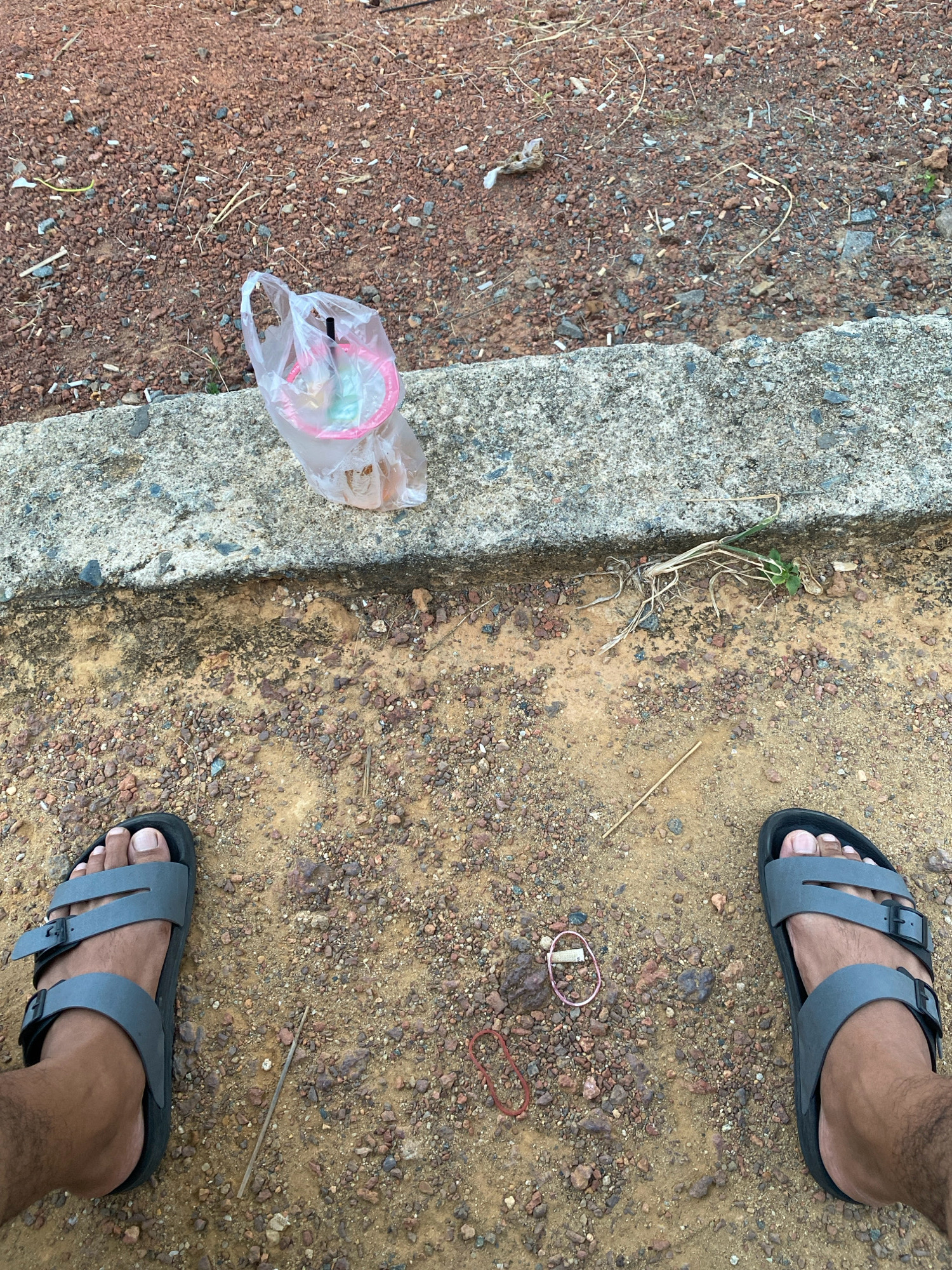 The image depicts a first-person perspective of a person wearing grey Birkenstock-style sandals, standing on a dirt and gravel surface. The ground is a mix of reddish-brown dirt and small pebbles. To the left, a concrete ledge runs horizontally across the frame. Resting on the ledge and partially in a clear plastic bag is a pink disposable cup with a straw, containing a beverage. Scattered on the dirt surface are a few dried plant stems and two rubber bands, one pink and one red. The background is a continuation of the reddish-brown dirt and gravel, suggesting an outdoor, possibly unpaved area. The presence of a concrete ledge and the general appearance of the ground suggest a roadside or pathway environment. The image does not provide specific visual cues to pinpoint the exact location within Tanjung Pinang, Indonesia, beyond it being an outdoor area with such ground cover and a concrete structure.