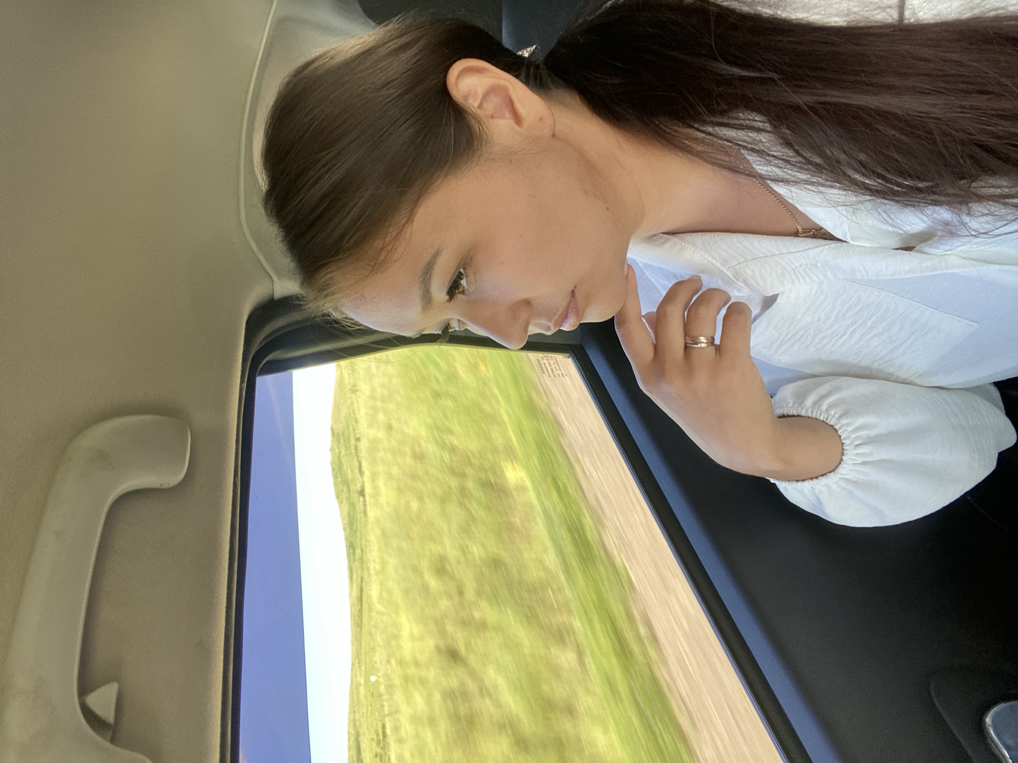 A young woman with dark hair pulled back in a ponytail sits in the back seat of a vehicle, looking out the window. She wears a white shirt with ruffles on the sleeves and a ring on her finger. The window reveals a blurred landscape of green hills and a blue sky, suggesting the vehicle is in motion. The interior of the car is visible, including a handle on the ceiling and the dark upholstery of the door. The scene appears to be in a rural area.