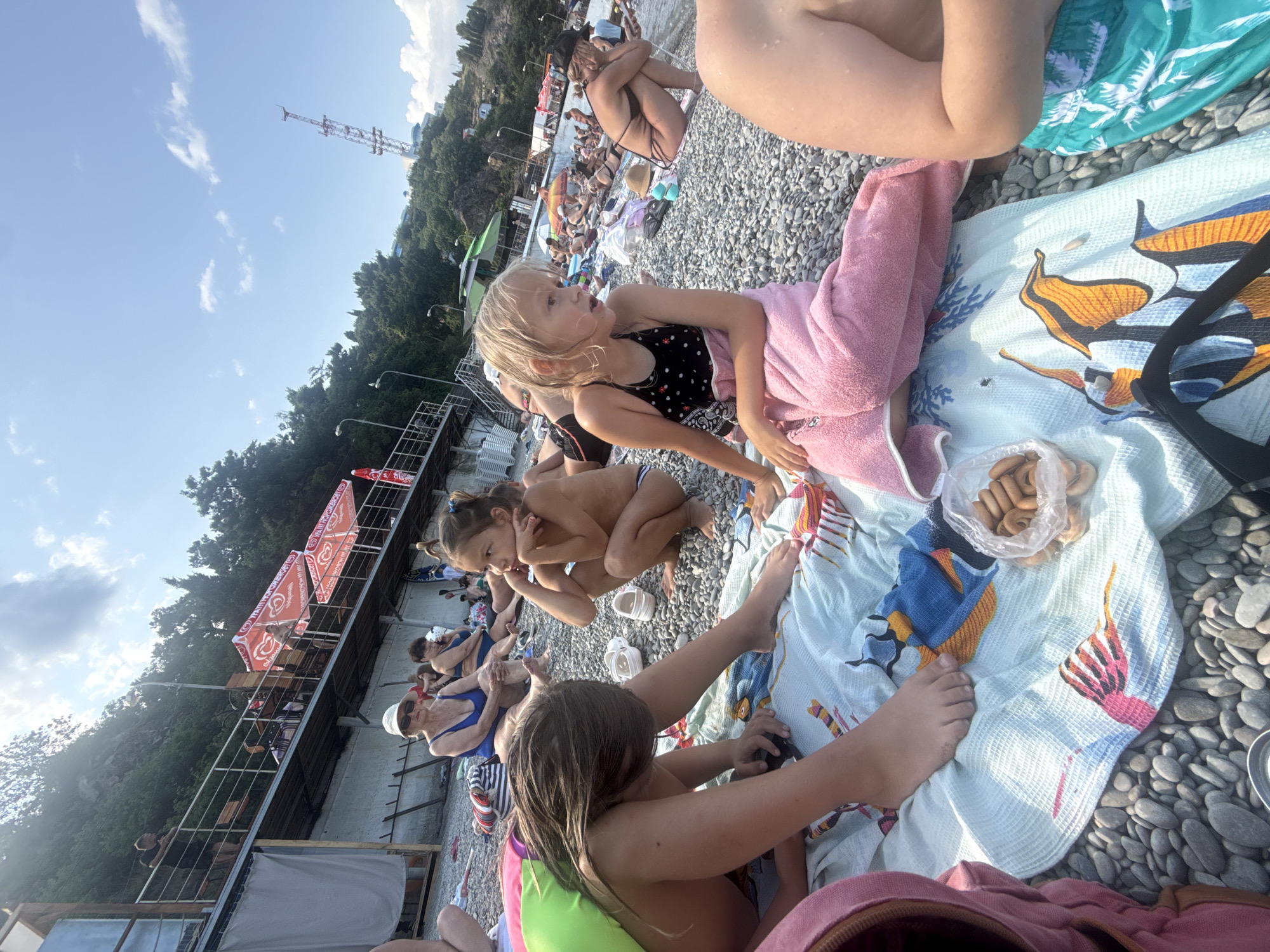 This is a wide-angle, eye-level shot of a crowded beach on a sunny day in Partenit, Ukraine. The foreground is dominated by a light blue towel with a fish pattern, spread on a pebble beach. Several children are seated on the towel. One child, with long brown hair, is wearing a green top and is leaning forward. Another child, with blonde hair and a black and white polka dot swimsuit, is sitting upright, wrapped in a pink towel. A plastic bag filled with small, round crackers is placed near the children.
In the middle ground, a woman with a white hat and sunglasses sits next to another woman in a blue swimsuit. Further back, many other beachgoers are visible, some sitting on towels and others walking. A wooden structure with red canopies, possibly a cafe or bar, is situated on a raised level behind the beach. Trees and a rocky incline form the backdrop, with a tall transmission tower visible in the distance on the hill. The sky is blue with scattered white clouds.