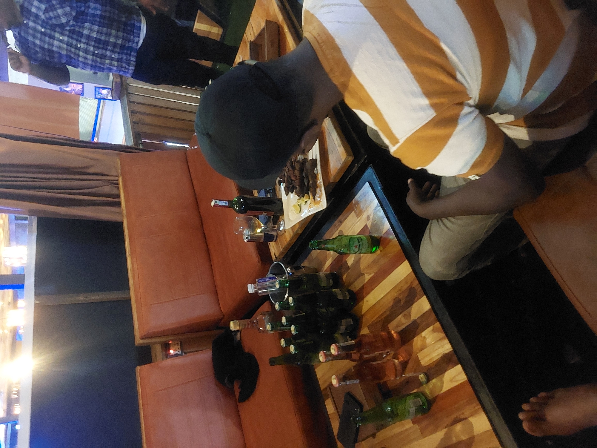 The image depicts an elevated outdoor dining area, likely in a restaurant or lounge, with a wooden plank table covered in numerous bottles of what appear to be alcoholic beverages. Two patrons are present, one wearing a blue and white plaid shirt standing in the background, and another wearing a white and brown striped shirt sitting at the table eating food. The table also features a bottle of red wine and a plate of grilled meat with lime slices. To the side of the table are orange cushioned seats. Based on the style of the establishment and its outdoor setting, it may be located in a tourist area within Dar es-Salaam, Tanzania.