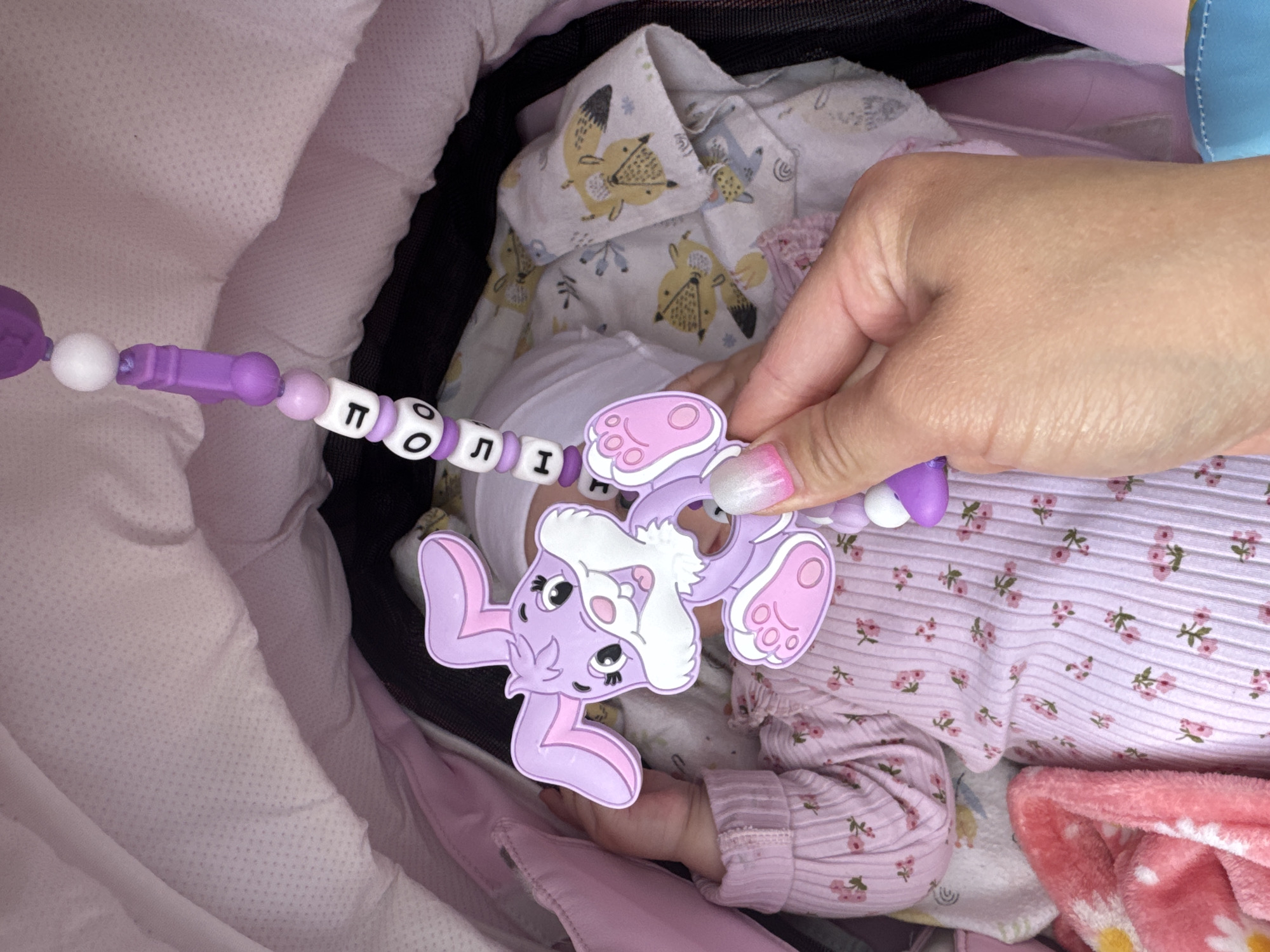 A close-up shot shows a hand holding a purple bunny-shaped teething toy. The toy is attached to a string of beads, some of which are white and purple, and others have black letters on them. The letters spell out "ПОЛІН", a name in Ukrainian. The toy and beads are positioned over a baby lying in a stroller. The baby is wearing a light pink outfit with a floral pattern and a white hat. The stroller interior is lined with pink fabric. In the background, a white blanket with a fox print is visible. The image appears to have been taken indoors during the daytime.