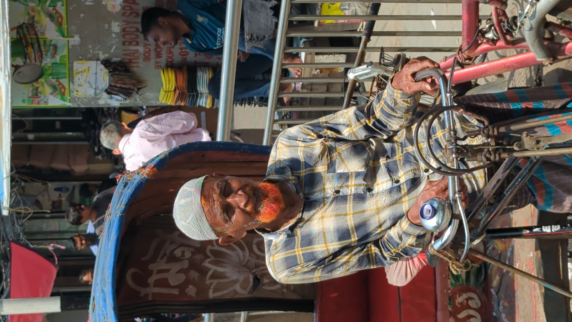 An elderly man with an orange-dyed beard and a white skullcap is seated on the driver's seat of a cycle rickshaw in Dhaka, Bangladesh. He is wearing a plaid shirt and gripping the handlebars. The rickshaw has a blue, decorated canopy. Behind him, partially visible, are other people, including a man in a pink shirt and another in a blue shirt. A "Thai Body Spa" sign is also visible in the background. The setting appears to be a busy urban street.