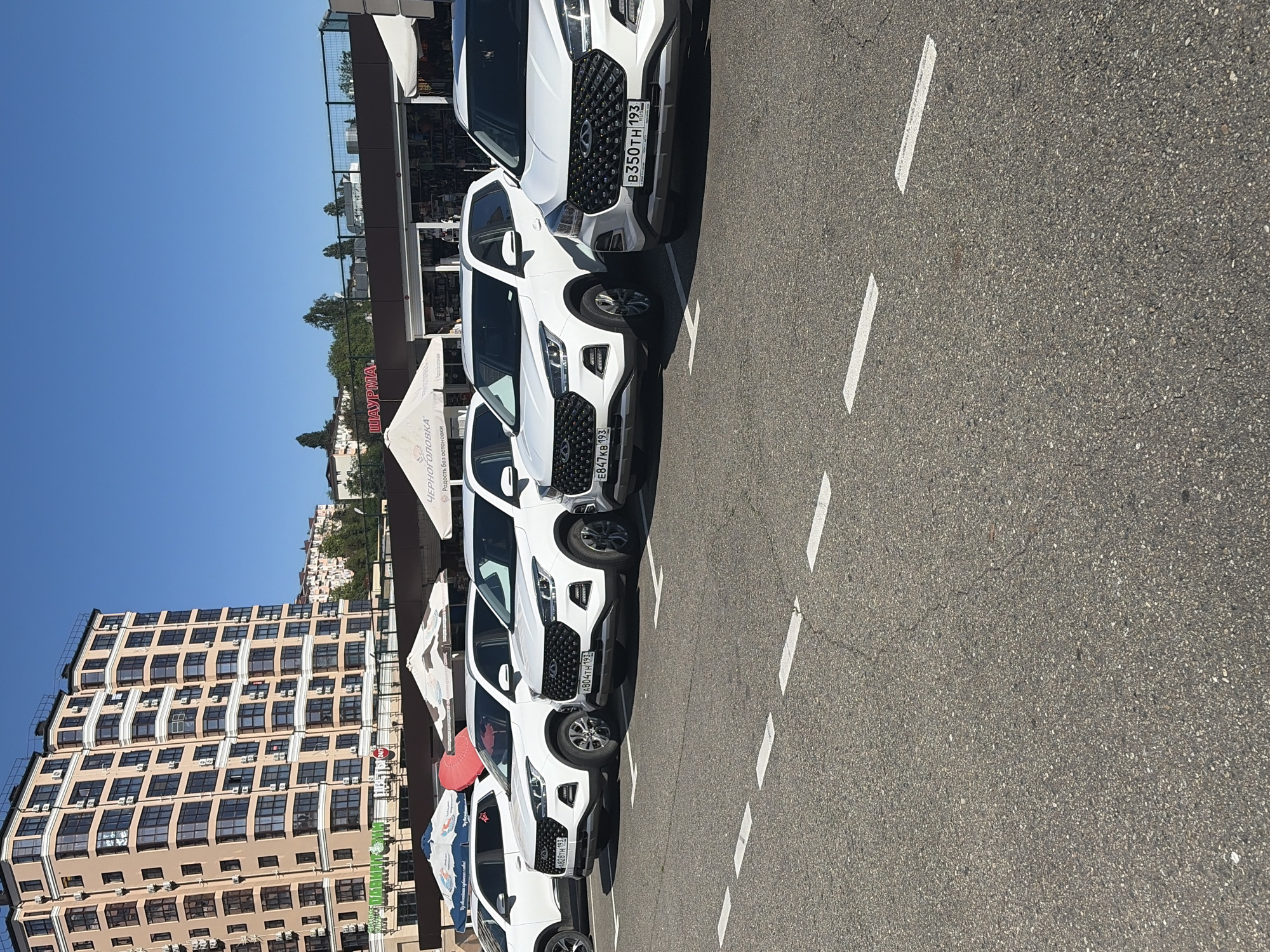 The image depicts a sunlit outdoor parking area with a row of five white SUVs, most likely Chery Tiggo 7 Pro models, parked side-by-side. The scene is set on a clear day, indicated by the bright blue sky and distinct shadows cast by the vehicles and buildings. The cars are parked on asphalt pavement, marked with white dashed lines separating parking spaces.
In the background, a multi-story building with numerous windows and balconies rises, showcasing a beige and dark brown facade. Many individual air conditioning units are visible attached to the building. To the right of the building, trees and other structures can be seen in the distance, suggesting a more developed area.
Several commercial tents or umbrellas are positioned near the parked cars and the building. Visible text on signs and tents includes "ШАУРМА" (Shawarma), "ЧЕРНОГОРОВКА" (Chernogorovka), and "Родость без остановки" (Joy without stopping). A sign on the building also reads "Медиахолдинг "Родина" (Mediaholding "Rodina").
The license plates on the vehicles are Russian, with the region code "193," which corresponds to Krasnodar Krai, indicating the location is consistent with Sochi, Russia. Specific plate numbers visible are B350TH 193, E847KB 193, A804TH 193, and A828TH 193. There is no explicit human activity or particular event taking place; the scene portrays a typical urban parking environment. No specific emotions are discernible. The time of day appears to be mid-day due to the high sun and strong lighting.