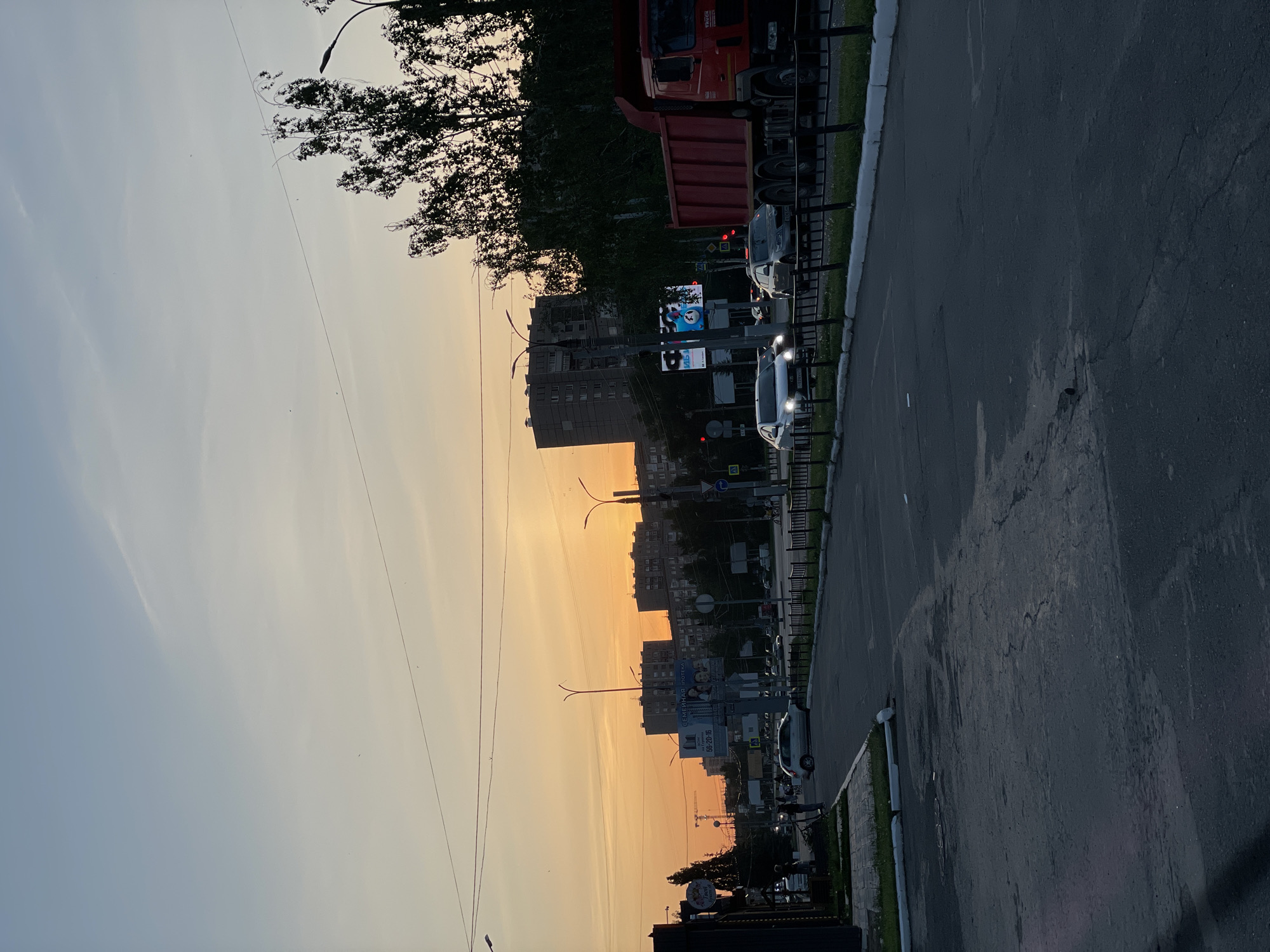 The image depicts a street scene in Izhevsk, Russia, at sunset. The sky transitions from a pale blue-gray to a vibrant orange-yellow. Tall buildings line the street, silhouetted against the sky, interspersed with streetlights and power lines. A road runs along the right side of the image, showing cracks and repairs. Several cars are on the road, and a red truck is parked on the roadside. A black metal fence separates the road from a narrow strip of green grass. A large billboard is visible along the street, displaying advertisements. Trees with dense foliage are also present.