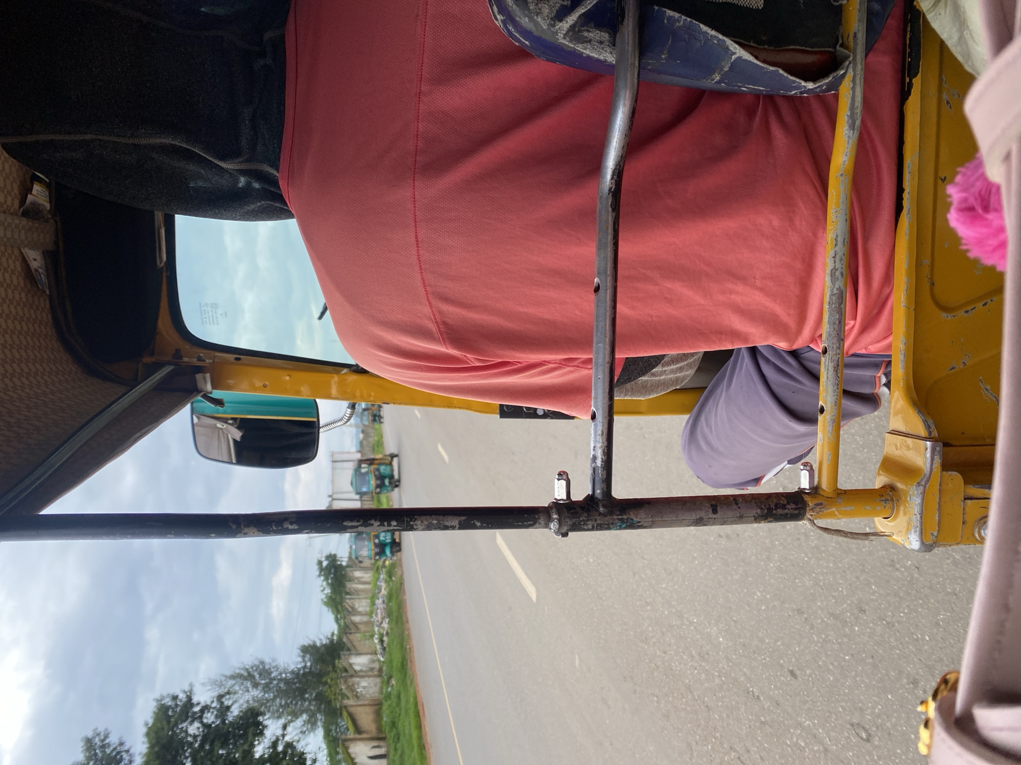 The image shows the interior of a yellow auto-rickshaw, likely a mode of transport in Rafin Par, Nigeria. The perspective is from a passenger, looking forward and slightly to the right.
The primary subject is the upper body of a person wearing a salmon-pink polo shirt. They are seated in the passenger area, which is upholstered in a patterned fabric. A blue and grey seat is visible behind them. The auto-rickshaw has a metal frame, parts of which are painted yellow and show signs of wear and tear. A side-view mirror, with a green casing, is mounted on the left side, reflecting a cloudy sky and other auto-rickshaws.
The scene appears to be a road, indicated by the asphalt surface visible below and to the right of the vehicle. Other auto-rickshaws are visible on the road in the distance, suggesting traffic or a common mode of transport in the area. The weather appears overcast, with a pale sky. The overall impression is of a daytime journey.
Notable details include a small pink fuzzy accessory hanging from the right side of the frame, possibly a keychain or decoration. The metal bars of the auto-rickshaw have a rough, weathered texture. No visible text is present in the image. The scene suggests an ordinary commute or travel within the specified location.
