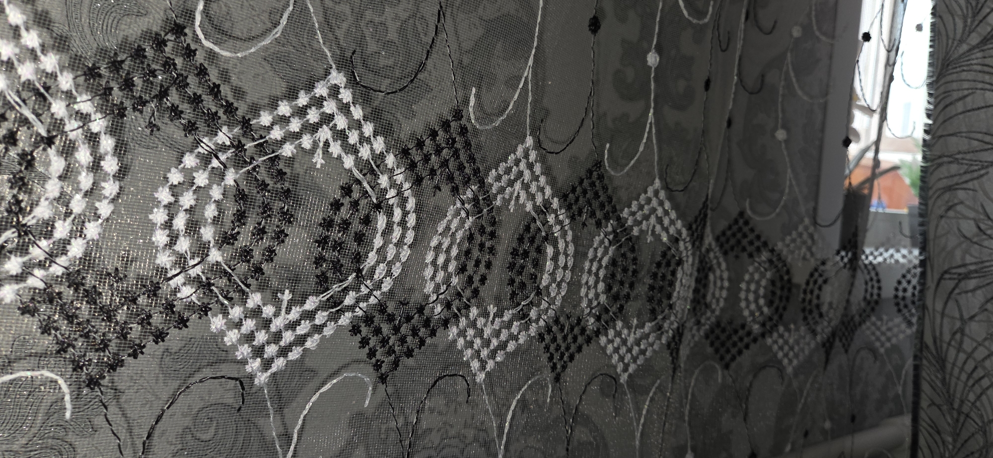 The image is a close-up, angled shot of a sheer black and white patterned curtain. The pattern consists of diamond shapes made of small stitched dots, alternating between black and white threads. Interspersed throughout the curtain are swirling, decorative white and black embroidered lines and small floral-like embellishments. The fabric is translucent, with a shimmering, metallic-like background pattern visible underneath the embroidery. To the right, a window is partially visible, showing a blurred outdoor scene with buildings and greenery, suggesting an urban or suburban environment. The curtain appears to be indoors, possibly in a residential setting. The lighting suggests it is daytime. There are no people or specific actions depicted.