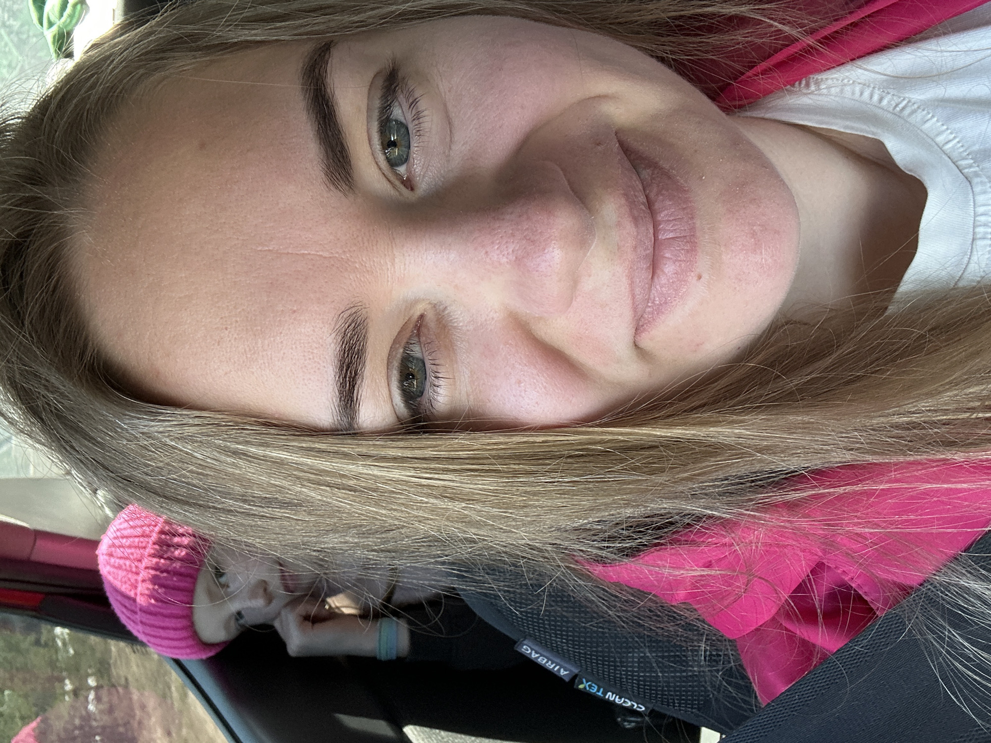 The image shows a close-up of a woman's face with light blond hair. She has fair skin, arched eyebrows, and grey-green eyes. The woman is wearing a white t-shirt and a pink hooded garment. In the bottom left corner of the frame, a second person, a young child wearing a pink beanie, is partially visible. The woman appears to be in a car, as indicated by the seat with an "AIRBAG CLEAN TEX" tag and a view of what appears to be an outdoor scene through the car window. The location is stated as being in the city of Фоки, Russia. The exact context or interaction is unclear, but the woman is facing the camera.