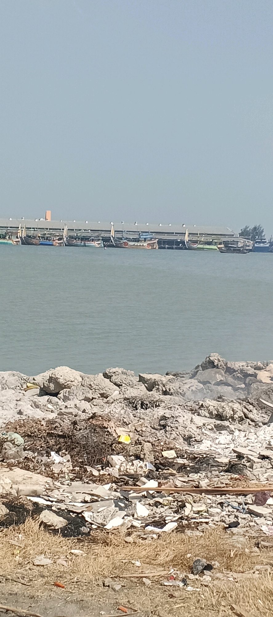 The image depicts a waterfront scene with a long building or pier in the background, lined with numerous boats of varying sizes and colors docked alongside it. The water is calm and a light blue-grey. In the foreground, the ground is covered with rocks, debris, and some dry vegetation. The debris appears to be a mix of building materials, plastics, and other discarded items, suggesting a somewhat neglected or polluted area. The sky is a clear, pale blue. The location is identified as Blimbing, Indonesia. There are no people visible in the image.