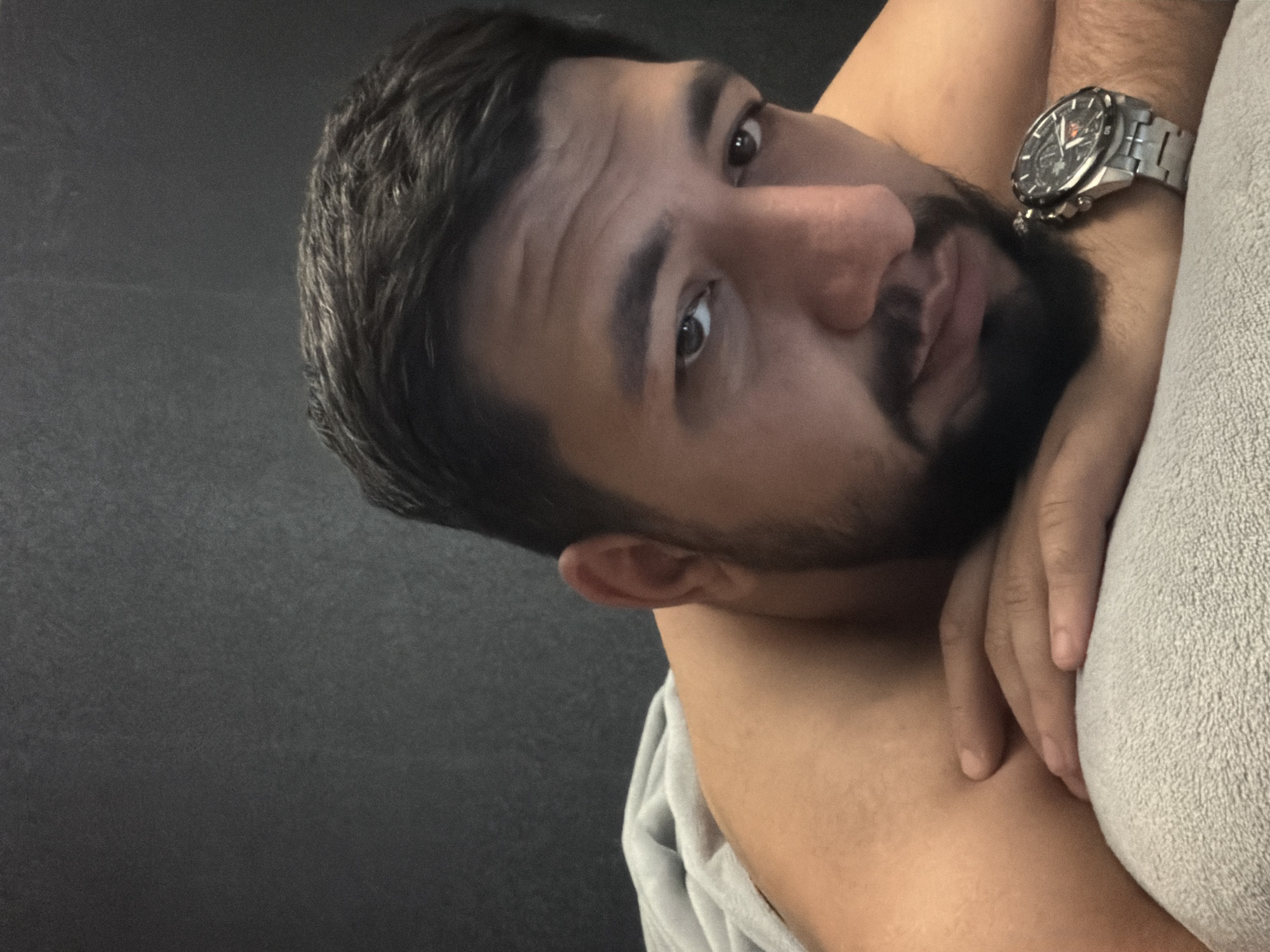 A close-up shot shows a man with brown hair and a beard looking at the camera. He is wearing a silver watch on his left wrist and appears to be lying down on a light grey towel. The background is a dark grey wall. The image does not contain information about the location in the city of Krasnoyarsk, Russia.