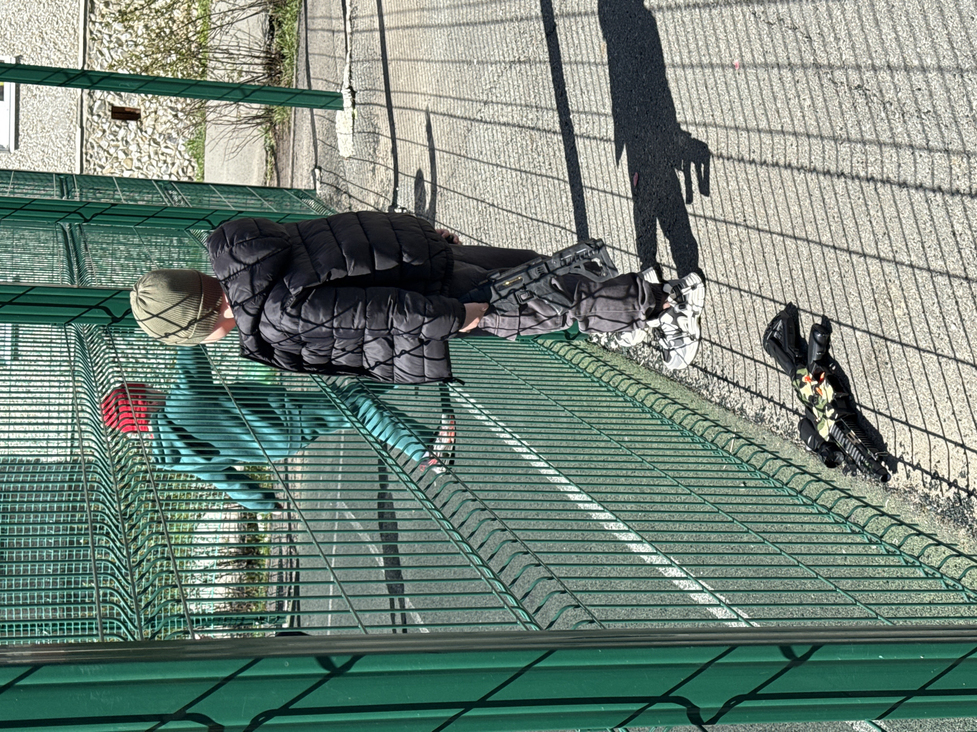 A male figure, wearing a dark quilted jacket and a light green beanie, stands by a green chain-link fence. Another person is visible in the background, wearing a light blue jacket with a red accent. Shadows are cast on the asphalt ground. A pair of shoes and a black bag rest on the ground. The image appears to be taken from an elevated perspective. The precise location in Yekaterinburg, Russia, is not discernible from the image itself.