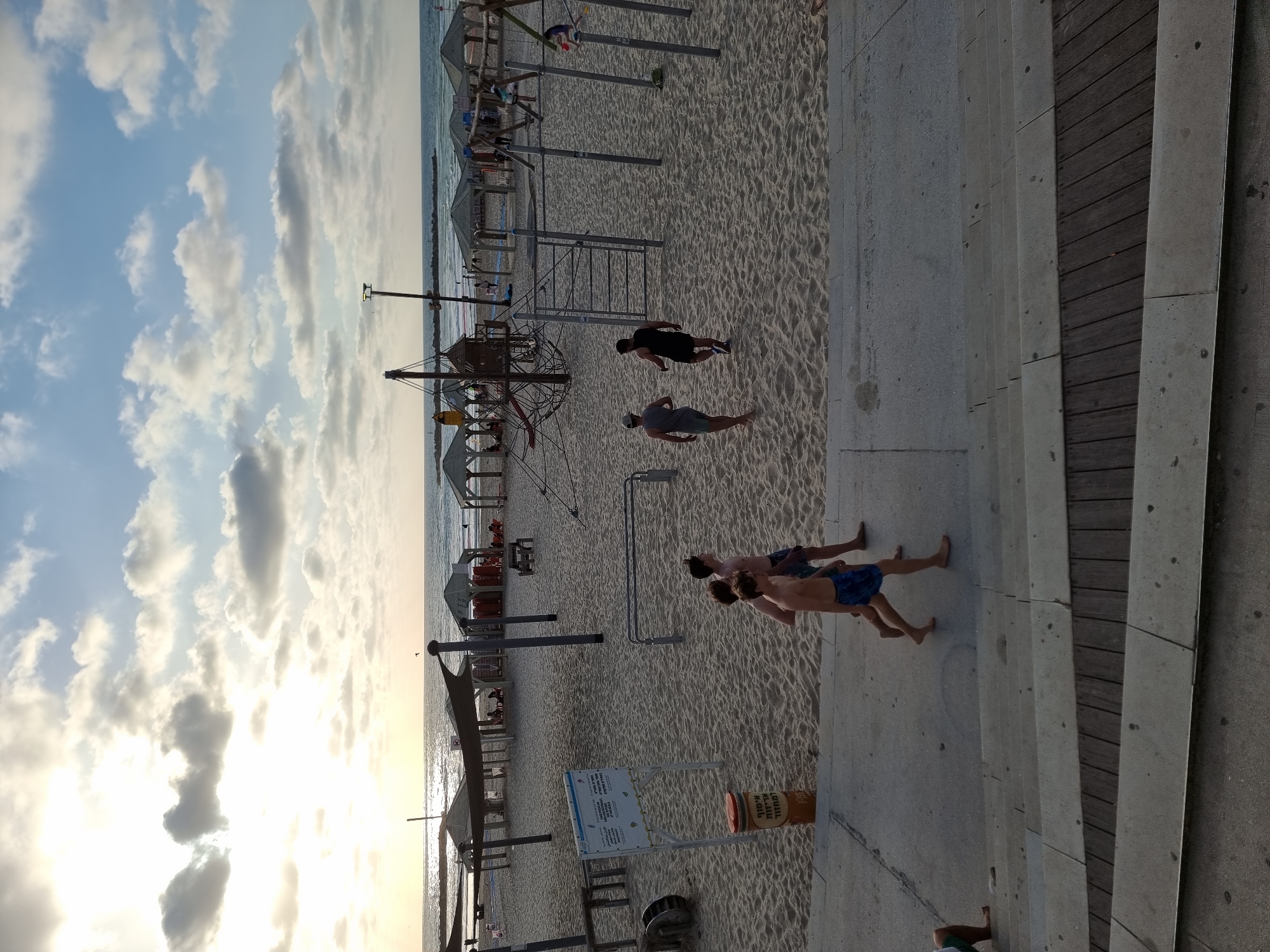 This image depicts a lively beach scene in Tel-Aviv, Israel, captured during the late afternoon or sunset, indicated by the low, bright sun and scattered clouds in the sky, which casts long shadows across the sand.
The scene is viewed from an elevated perspective, looking down onto a concrete boardwalk and steps on the right, transitioning to a wide expanse of sandy beach in the middle, and the Mediterranean Sea on the left horizon.
Several people are present, enjoying the beach. On the concrete steps, three men walk barefoot, two shirtless (one in blue swim trunks, another in light blue swim trunks), and one in a dark t-shirt and shorts. Their relaxed posture suggests leisure activity. On the sandy beach, two men walk past calisthenics equipment. Further down the beach, near the water, multiple beach umbrellas and loungers are set up, and a large pirate ship-themed playground structure is visible. The sea appears calm, with reflections of the sun shimmering on its surface.
Notable details include a sign with visible text primarily in Hebrew, situated near the boardwalk on the sand. Adjacent to it is a large brown cylindrical bin, also featuring Hebrew text that appears to say "תוספת תוסף" (additive). The overall atmosphere is serene and typical of a bustling beach destination.