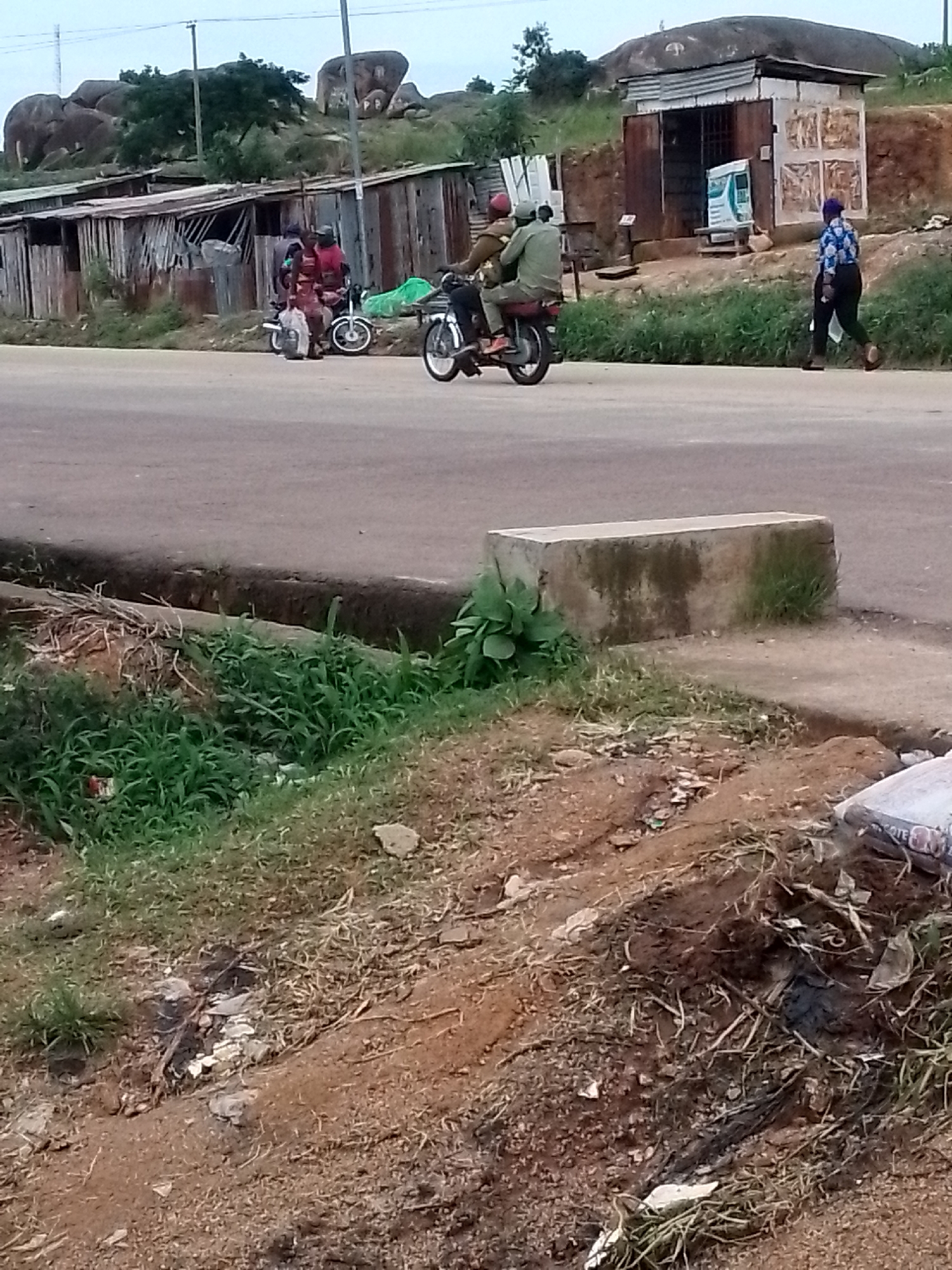 The image depicts a street scene in Jos, Nigeria, featuring a road and its surroundings. In the mid-ground, two motorcycles are visible. The first, on the left, carries two people, one in a red shirt and another behind, both facing forward. The second motorcycle, to the right of the first, transports two individuals clad in green, also facing forward. A pedestrian, wearing a blue and white patterned top and dark trousers, walks on the right side of the road, moving away from the viewer. Behind the motorcycles, several structures are present, including a long, weathered building constructed with corrugated metal and wood, and a smaller kiosk-like structure with similar materials. In the background, a hilly landscape with large rock formations and vegetation is visible under an overcast sky. The foreground shows a patch of disturbed earth with dry vegetation and debris, bordering the road.