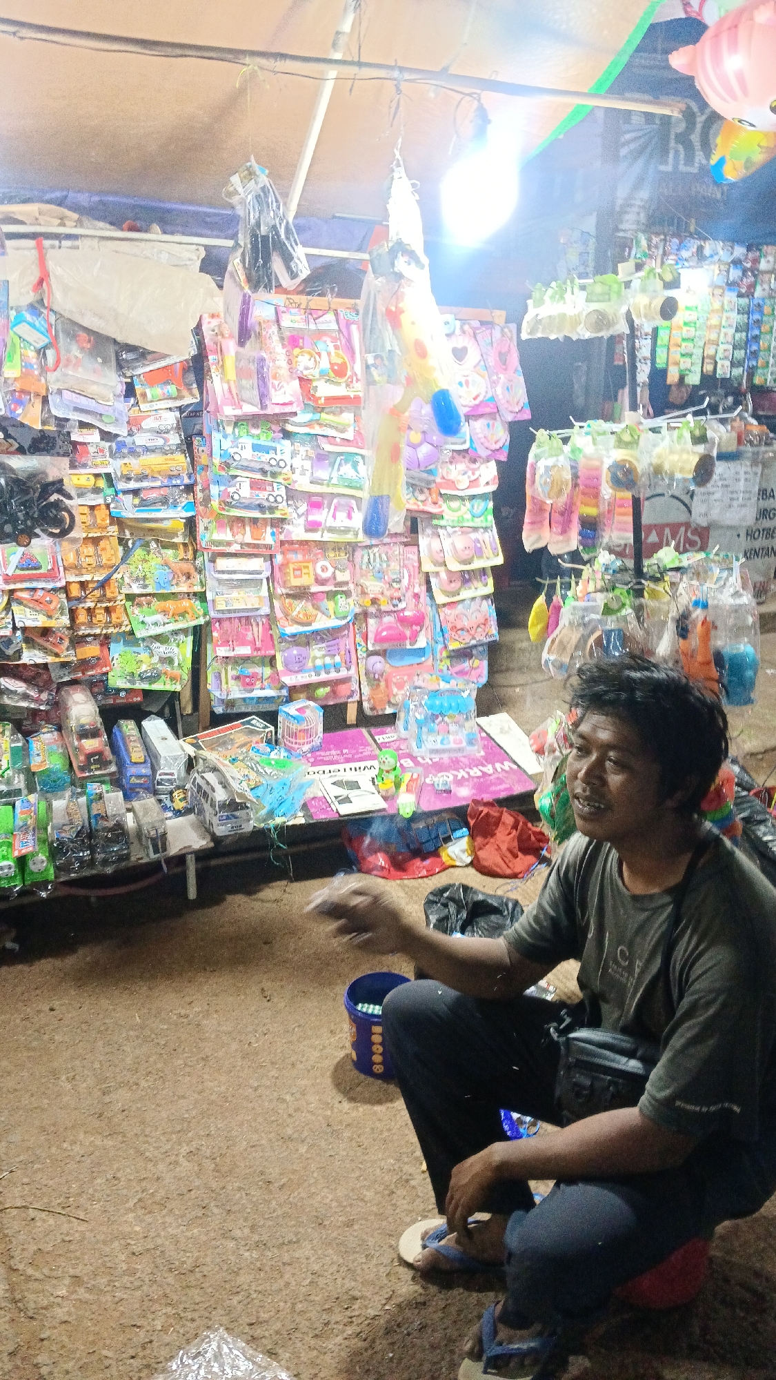 In Tajurhalang, Indonesia, a man with dark hair and fair skin sits on a red object on the ground. He is wearing a dark gray t-shirt, black pants, and blue flip-flops. He has a black bag slung across his chest. To his right, a blue bucket is visible. In front of him, a stall is laden with various children's toys and novelty items, including toy cars, stuffed animals, and colorful packets of goods. The stall is illuminated by a bright light bulb hanging overhead, casting a glow on the merchandise and the surrounding dirt ground. Behind the stall, more items are displayed, and a pink inflatable toy is partially visible. The scene suggests an outdoor market or a street vendor setup.