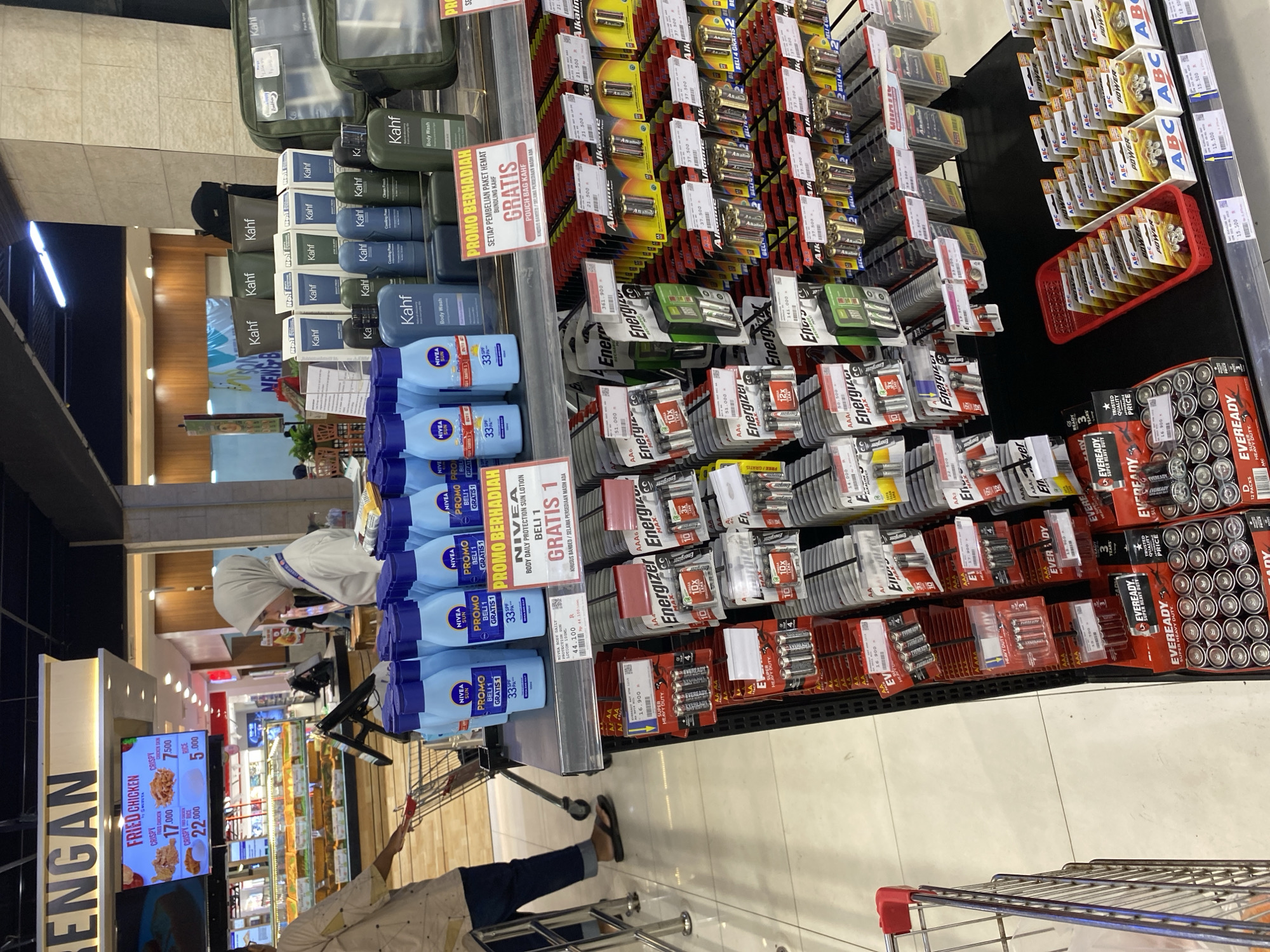 The image depicts a retail store, likely a supermarket or hypermarket, with various consumer goods displayed on shelves. In the foreground, rows of batteries, primarily AA and AAA sizes, from brands like Energizer and Eveready, are stacked. To the left, several blue bottles of Nivea body lotion are on display, with promotional signage indicating a "Buy 1 Get 1 Free" offer. Further back, a selection of Kahf brand toiletries, including body wash, is visible. A person wearing a hijab and a light-colored uniform, possibly an employee, is attending to a display of Kahf products. In the background, a food display, possibly for fried chicken, is visible, with prices listed in Indonesian Rupiah (Rp). The overall environment appears to be a well-lit commercial space with tiled flooring. While the image does not explicitly state the location, given the Indonesian currency and product branding, it is plausible that the store is located in Indonesia, and potentially in a city like Banda Aceh, Indonesia, as suggested by the prompt.