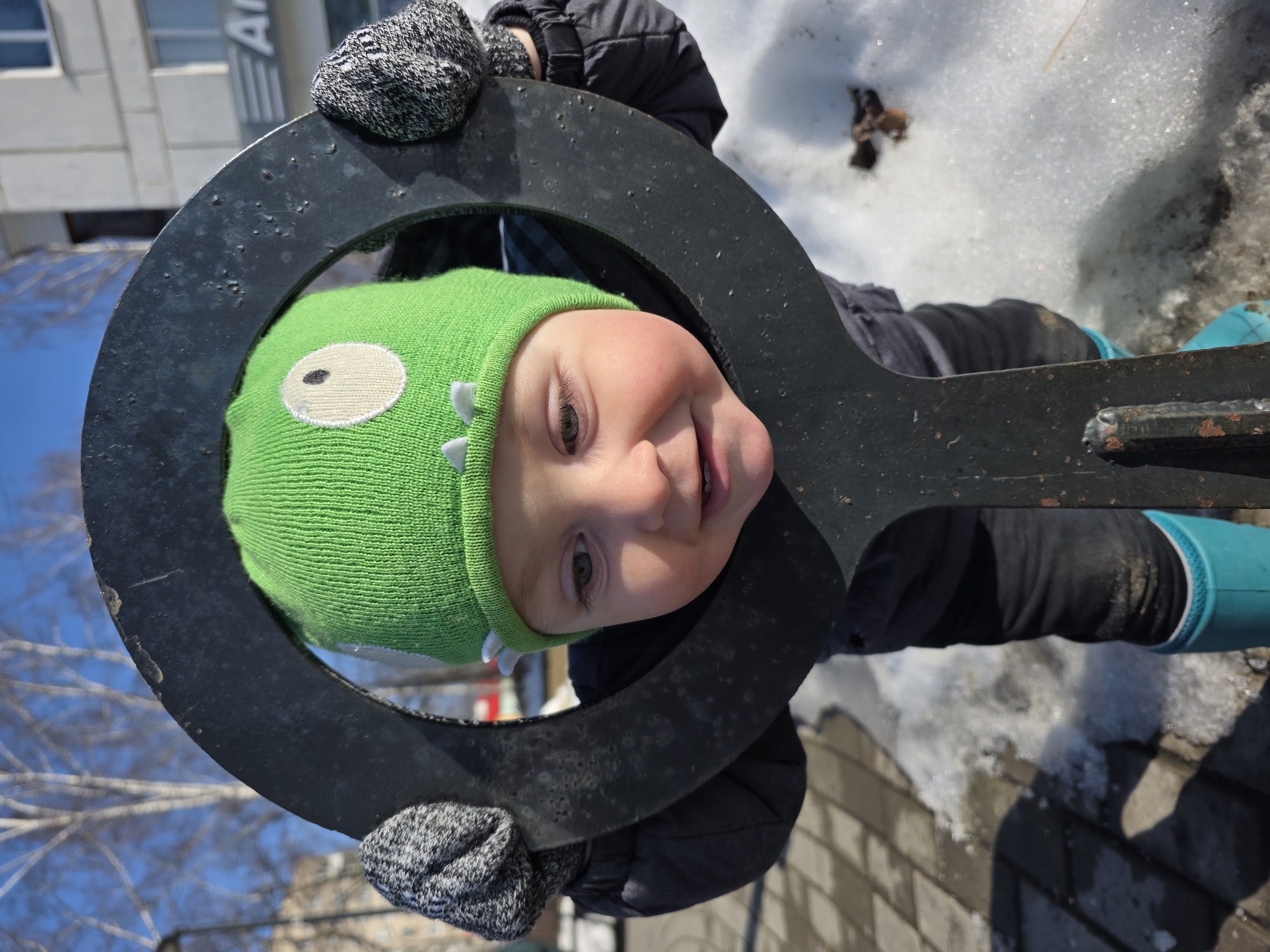 This image depicts a young child in an outdoor urban setting within the city of Chaikovsky, Russia. The child, appearing to be a toddler, is centered in the frame, wearing a bright green knitted beanie designed to resemble a monster or dinosaur with white eyes and small white spikes, a dark winter jacket, grey patterned mittens, dark trousers, and turquoise rubber boots. The child's face, framed within a large, dark-colored, worn metal ring of what appears to be a piece of playground equipment or a decorative structure, shows light-colored eyes and a slight smile. The child grips the metal ring with both gloved hands.
The ground beneath the child consists of light-colored paving stones interspersed with patches of melting snow and ice, indicating a late winter or early spring season. In the background, bare deciduous trees are visible against a clear blue sky. Behind the trees, a multi-story, light-colored building with visible windows features a partial sign displaying the Cyrillic letter "A". The scene is well-lit by direct sunlight, casting shadows.