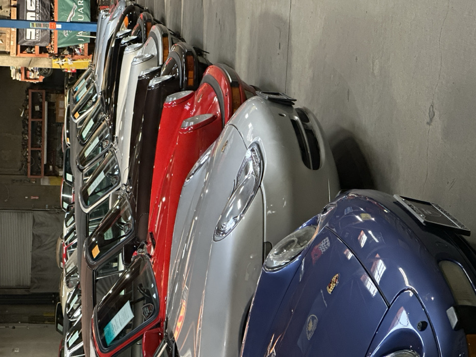 A row of cars, predominantly vintage models, are parked closely together in an indoor space, possibly a warehouse or garage. The vehicles display a range of colors including blue, silver, red, brown, and white. The image is shot at an angle, focusing on the front portions of the cars, highlighting their headlights and hoods. A shelf with a Jaguar banner is visible in the background, suggesting the location might be a car dealership or a storage facility specializing in classic automobiles.
