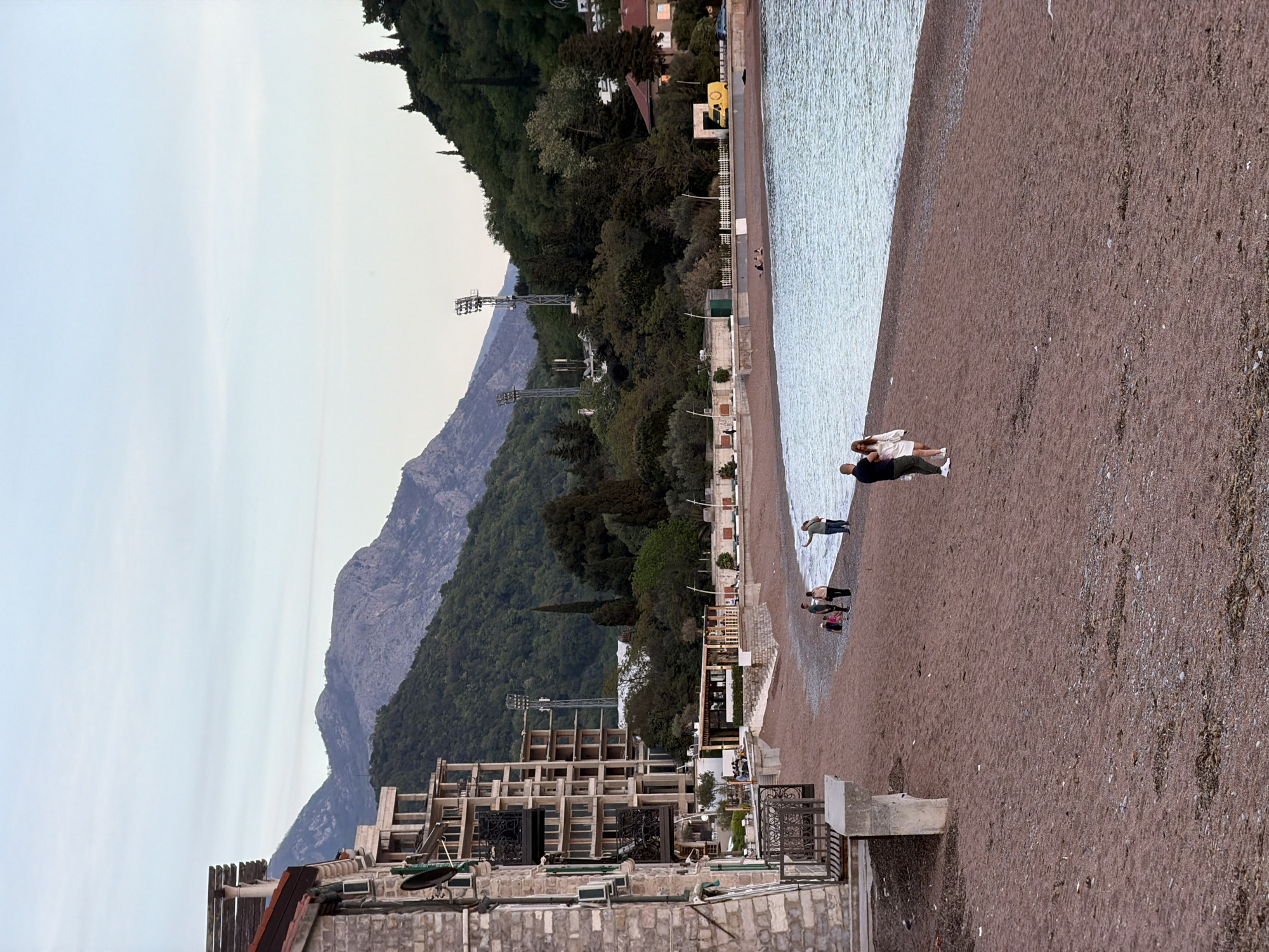 The image shows a beach in Petrovac centar, Montenegro. The beach is sandy, with some pebbles visible. The water is light blue and relatively calm. There are a few people on the beach, including a couple standing near the water's edge and a family further up the beach. The background includes a mountain range, a forested hillside, buildings and some scaffolding. The sky is light blue. The picture seems to have been taken around dusk or dawn.
