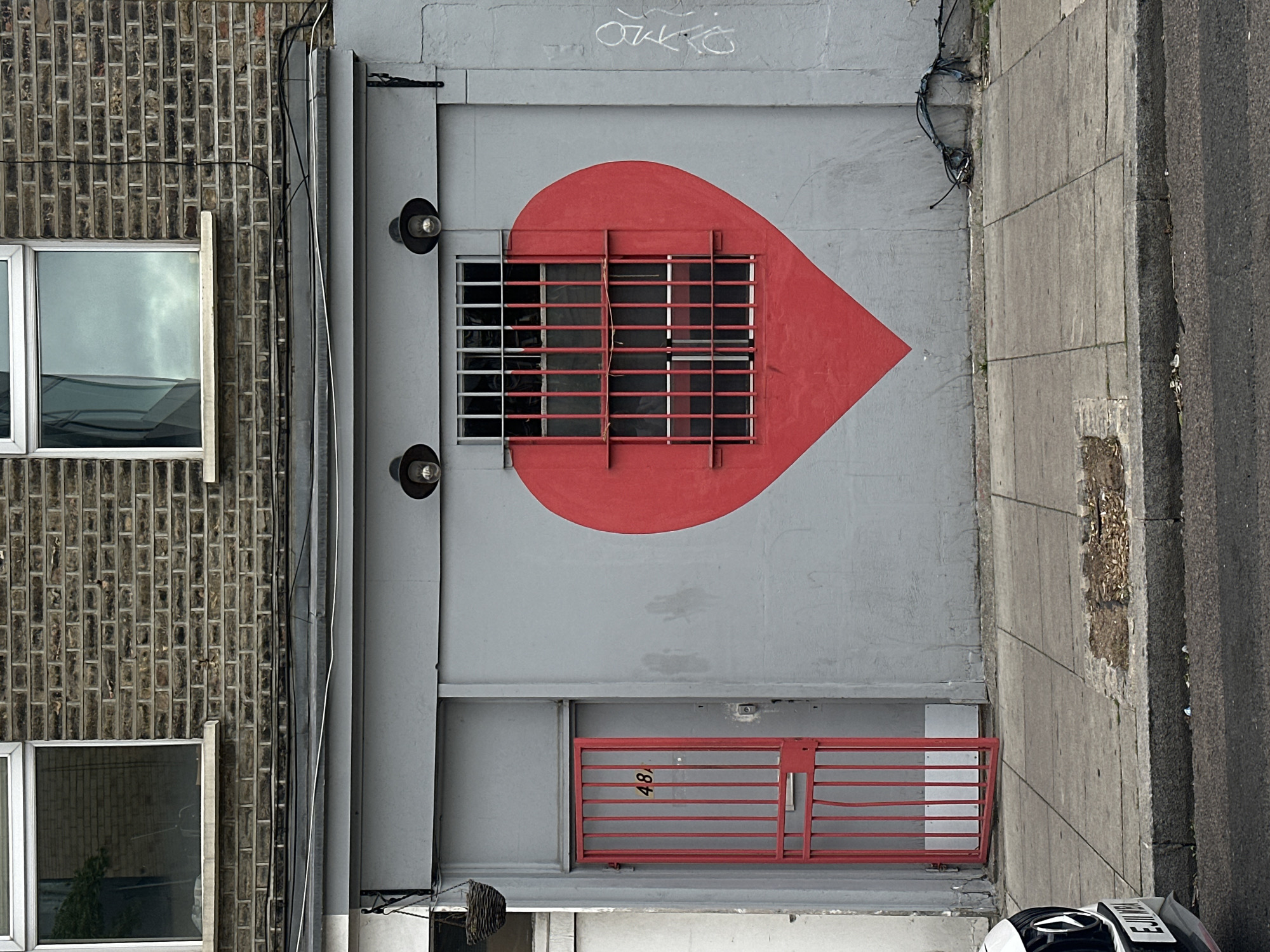 The image shows an exterior wall of a building in London, United Kingdom, featuring a large red heart painted on a grey wall. The heart is partially obscured by a red metal grille covering a window. Below the heart, another red metal gate is visible, seemingly covering a small opening. The number "48" is faintly visible on the gate. Two circular light fixtures flank the heart near the top. A brick building is attached to the left of the grey wall, with two windows visible. A sidewalk runs along the right side of the grey wall, adjacent to the street. A partially visible motorcycle with a license plate is parked near the sidewalk. Graffiti is present on the grey wall above the heart.