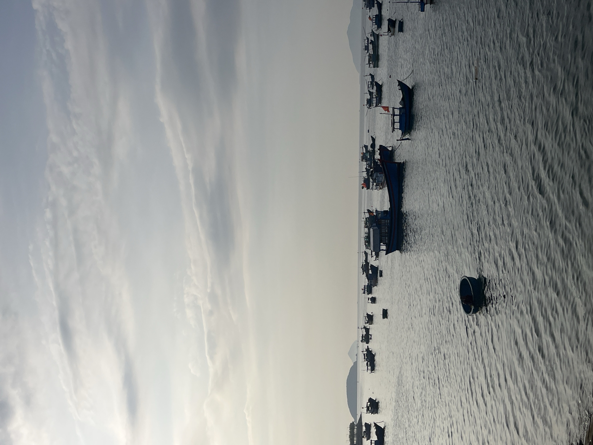 The image depicts a marine scene in Nha Trang Ward, Vietnam, with numerous small boats and ships scattered across the water. The sky is overcast with high-altitude clouds. Several fishing boats of different sizes are visible, some with small red flags. The water has a textured surface, indicating slight waves. A distinct round boat is centrally positioned. In the distance, there is a faint outline of land or islands. No people are clearly visible in the image.