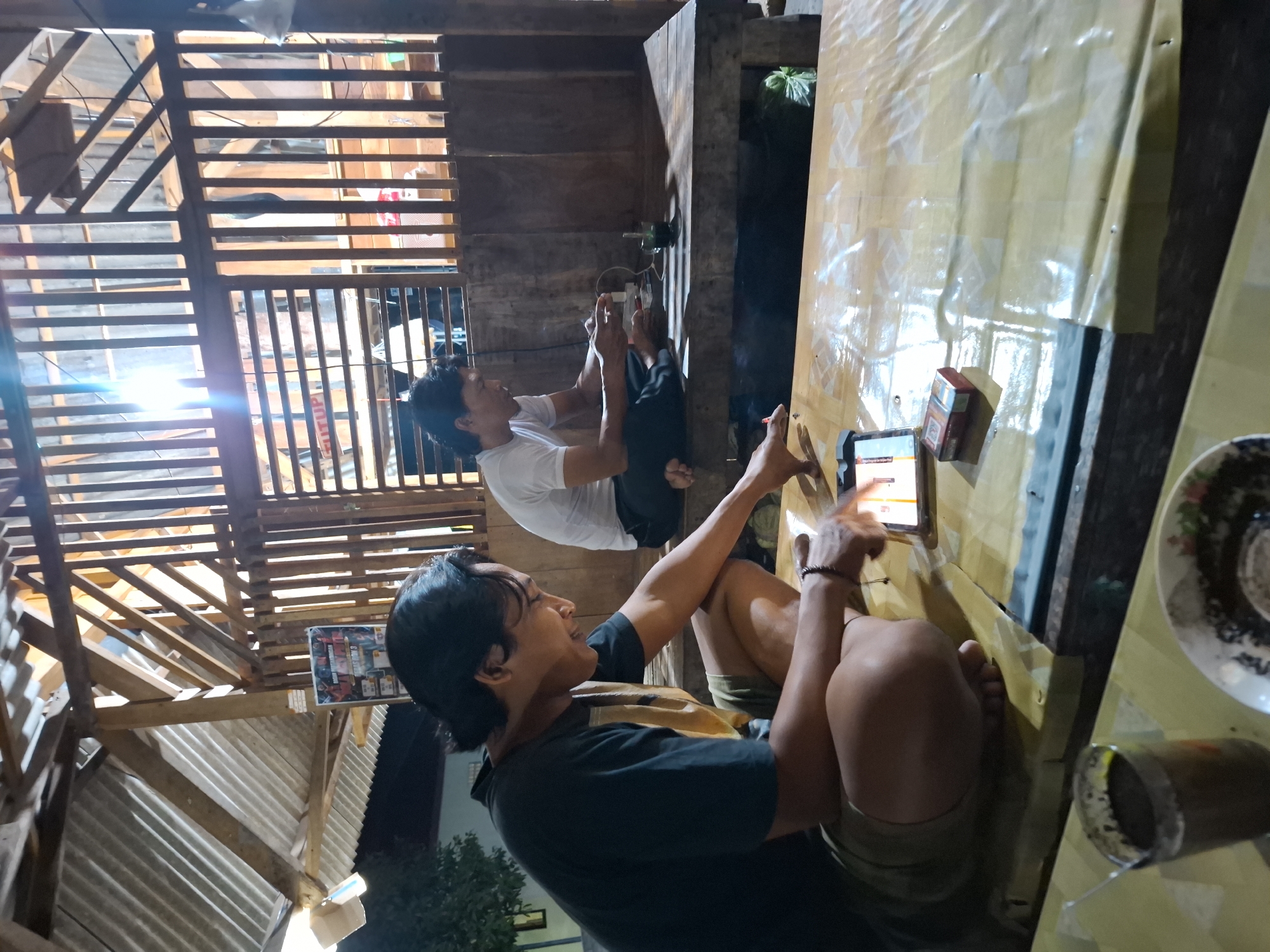 Two men are sitting on the floor of an open-air structure with a wooden lattice wall. The man on the left, wearing a dark t-shirt and shorts, is looking down at a tablet and appears to be interacting with it. In front of him on a table covered with a yellow tarp, there is a pack of cigarettes and the tablet. A glass of dark liquid is to his left. The man on the right, wearing a white t-shirt and dark pants, is also looking down at something in his hands, possibly a phone, and appears to be smoking. He is sitting on a wooden bench. The structure has a corrugated metal roof supported by wooden beams. The environment appears to be a casual outdoor setting, possibly a cafe or gathering spot. The scene is lit by a bright overhead light.