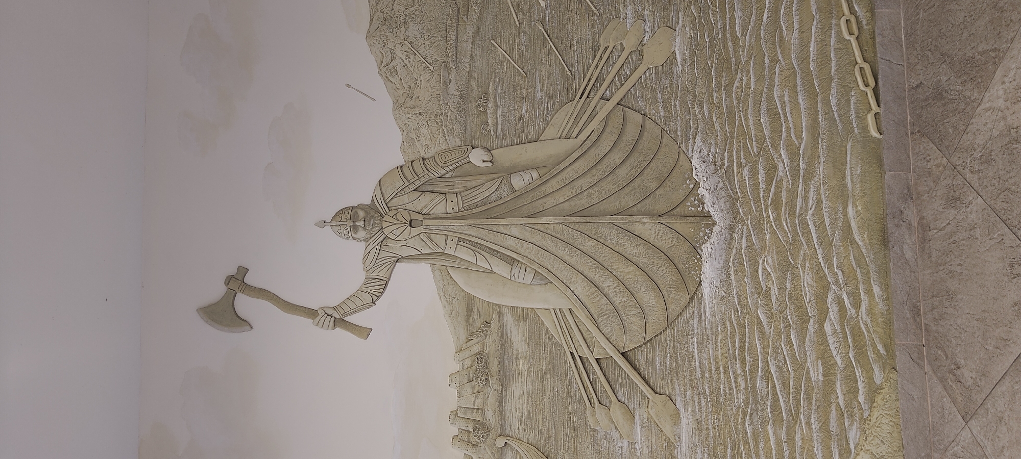 The image depicts a bas-relief carving on a wall, possibly in Khujand, Tajikistan. The carving features a Viking warrior in a boat, holding an axe aloft. The warrior is dressed in ornate armor and a helmet, with a beard and stern expression. The boat is a longship with oars visible. In the background, there is a fortified settlement on a hill, a body of water, and mountainous terrain. The carving is detailed, with textures representing water, clouds, and stone. The overall color palette is monochromatic, with shades of beige and off-white. The floor in the foreground appears to be tiled.
