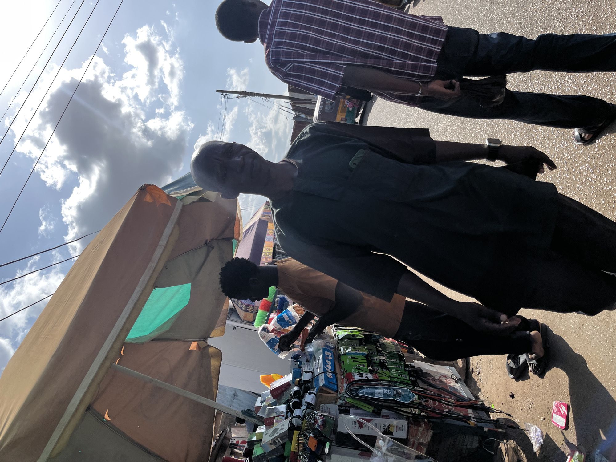 The image depicts a street scene in Gura Top, Nigeria, under a bright, partly cloudy sky. In the foreground, a bald Black man wearing a dark green, collared shirt is the primary subject, facing the camera with a neutral expression. To his left, another Black man, partially visible, wears a dark shirt. To the right of the main subject, a Black man in a maroon and white plaid shirt walks away from the camera, holding a dark bundle in his right hand.
Behind the main subject, a stall is set up under a large, tan awning. The stall is laden with various items, predominantly electronics and accessories such as chargers, cables, and headphones, displayed on shelves and racks. A younger Black man, wearing an orange t-shirt, is attending to the stall, holding what appears to be a broom and a tablet. His attention is focused on the merchandise.
The ground is paved and appears wet in patches, reflecting the sunlight. Debris, including a pink object, is scattered on the ground near the stall. In the background, a light-colored auto-rickshaw is visible, along with corrugated metal structures and utility poles with numerous wires. The overall impression is that of a bustling market environment.
