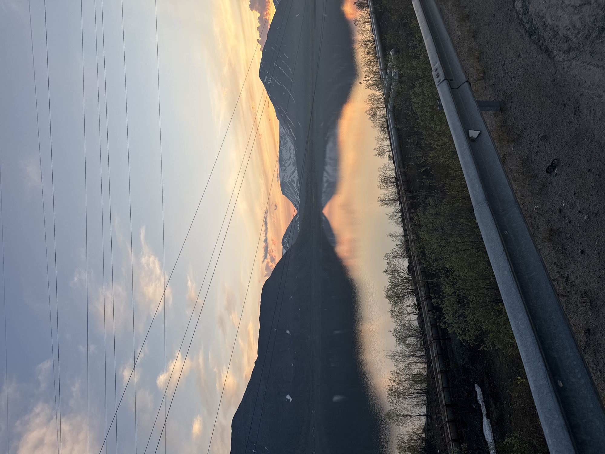 A wide-angle landscape photograph depicts a mountainous lakeside scene at either sunrise or sunset, characterized by a serene body of water reflecting warm sky colors and dark mountain silhouettes. The overhead sky transitions from a light blue at the zenith to vibrant orange and pink hues near the horizon, populated by scattered, wispy clouds. Several dark, taut power lines traverse the upper portion of the frame diagonally, descending from left to right.
In the background, a formidable range of dark mountains, partially covered with patches of white snow, rises majestically, its contours sharply reflected in the still water of the lake. The mountain on the far right exhibits more pronounced snow lines. The midground is dominated by a large, placid lake, which acts as a mirror, perfectly reflecting the sky's warm tones and the dark forms of the mountains, creating a striking symmetrical composition.
The foreground features a galvanized steel guardrail extending diagonally from the lower right corner, partially obscuring the view. A small white triangular reflector is attached to one of its vertical posts. Below and behind the guardrail, the ground is a mix of dark earth and sparse, light green deciduous vegetation and bushes. Directly behind this vegetation, a series of thick, grey industrial pipelines runs horizontally along the base of the mountains, parallel to the lake shore, indicative of significant infrastructure. A patch of residual snow is visible on the ground to the left of the guardrail.
This scene is consistent with the geographical and industrial characteristics of Kirovsk, Russia, located within the Khibiny Mountains, known for their mining operations and associated infrastructure, and large bodies of water that reflect the dramatic sky conditions of high-latitude sunrises or sunsets. No people, active subjects, or specific interactions are visible within the frame. The environment is a subarctic or boreal mountainous landscape.