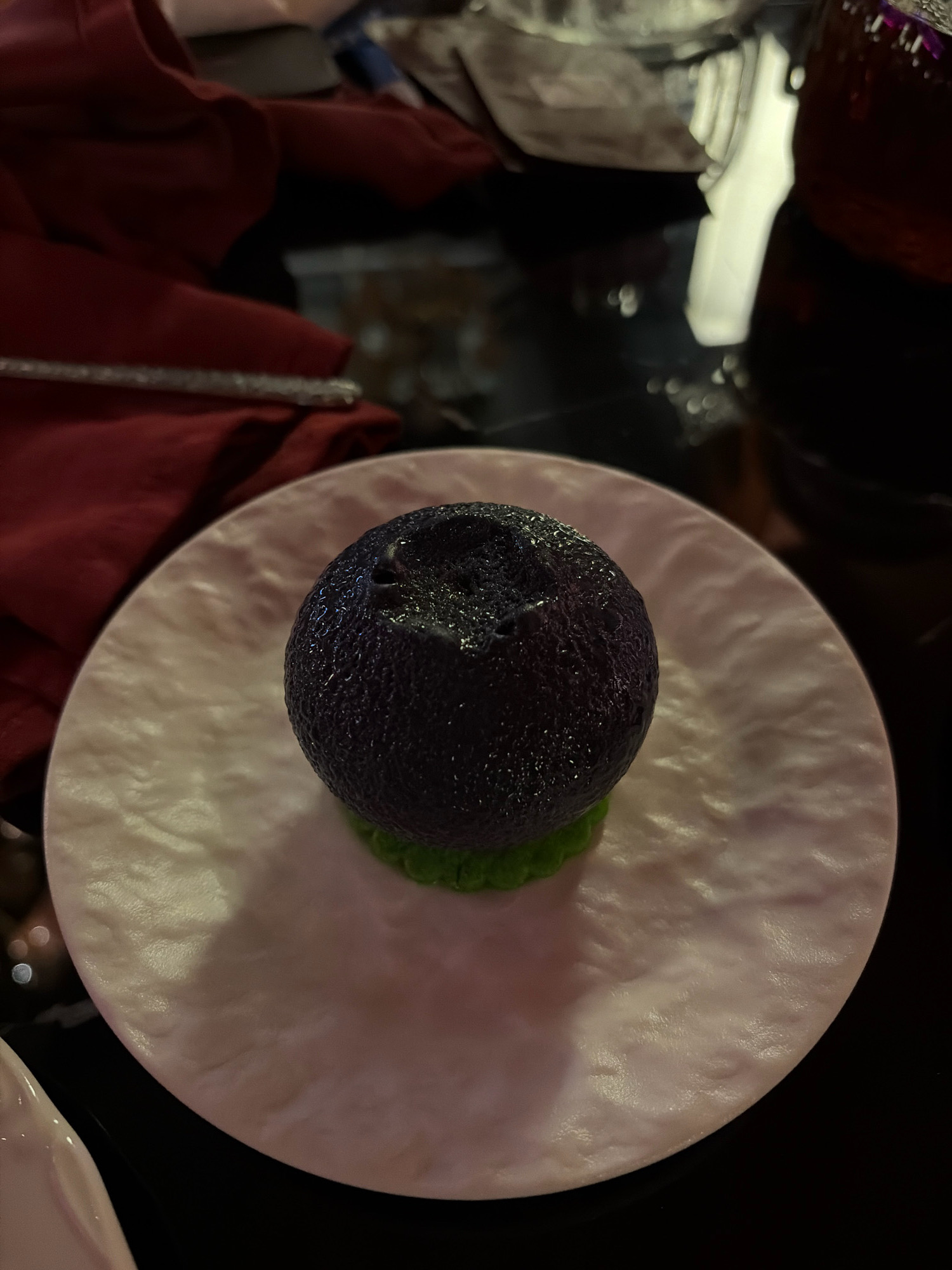 The image shows a close-up view of a dessert on a pink, textured plate. The dessert consists of a dark purple, spherical element with a textured surface, sitting on a small mound of bright green puree or mousse. The plate is placed on a dark, reflective surface, possibly a table. In the background, a red cloth is visible, along with hints of silverware and a glass. The lighting is dim, creating some glare and reflections. The scene appears to be indoors, possibly in a restaurant. There is no indication of the city of Astana, Kazakhstan.