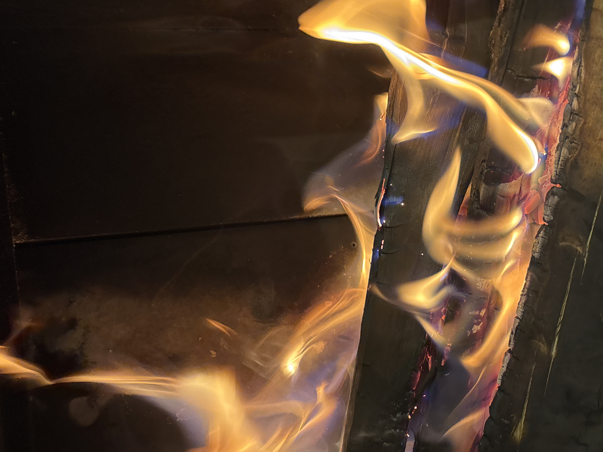 The image depicts a close-up view of a fire burning within a fireplace or wood-burning stove. Logs are visible at the bottom of the frame, with flames consuming them and rising upwards. The flames are a vibrant mix of orange, yellow, and some blue hues, suggesting high temperatures. The background appears to be dark and somewhat textured, possibly the interior of the fireplace or a dark wall. The scene is devoid of any people or specific identifiable landmarks, and therefore, its location in Zvenigorod, Russia, cannot be determined from the visual information provided. The image captures the dynamic nature of fire with its fluid shapes and intense colors.
