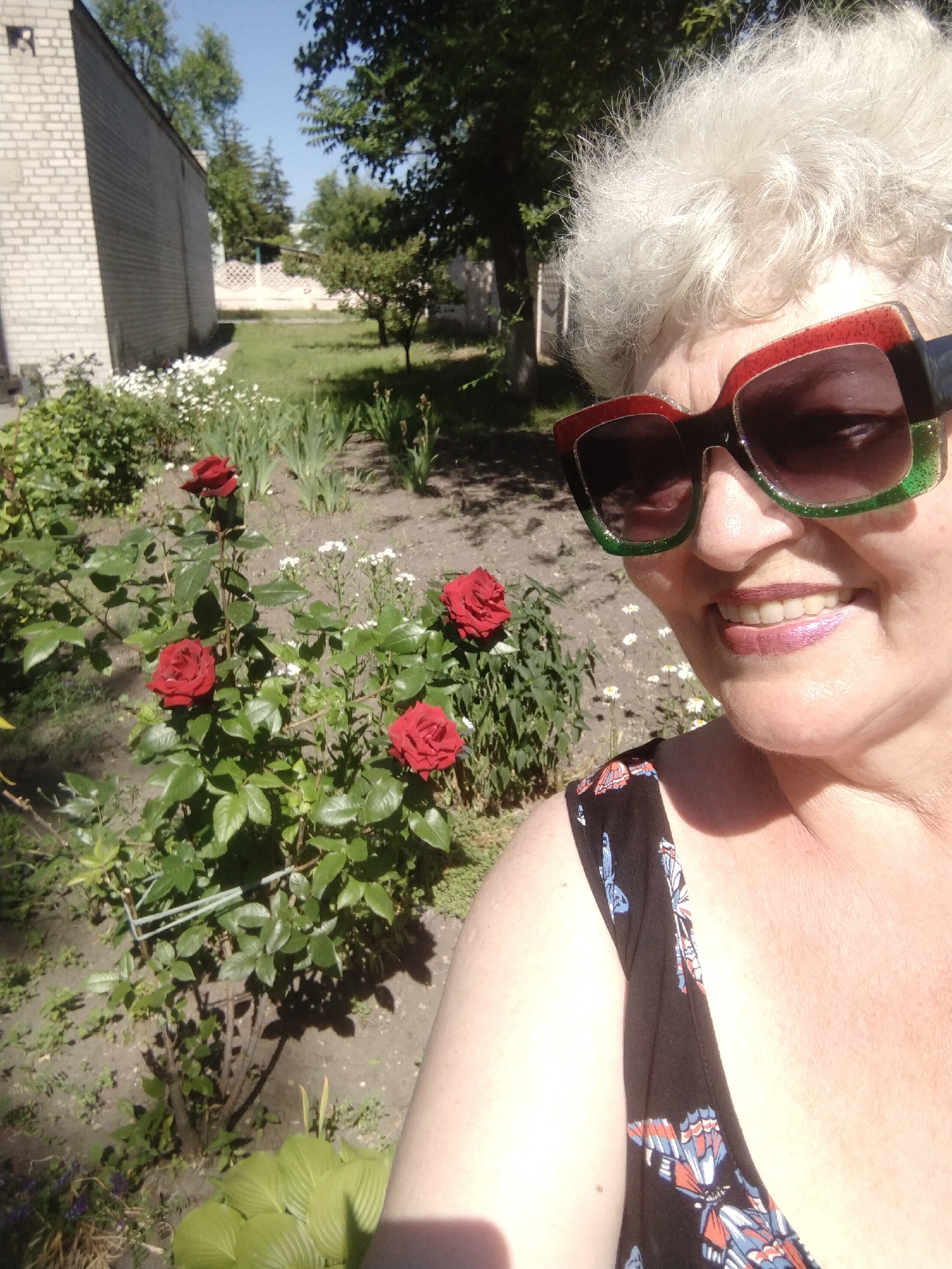 A medium shot captures a woman taking a selfie outdoors in front of a garden. She is wearing large, colorful sunglasses with red and green rims, and a black dress with a butterfly print. Her face is angled towards the camera, and she is smiling. In the background, there are rose bushes with red roses, green foliage, and other small white flowers. A brick building is visible to the left, and trees and a clear blue sky are in the distance. The scene appears to be a residential area in Horishni Plavni, Ukraine, likely a private garden or backyard on a sunny day.