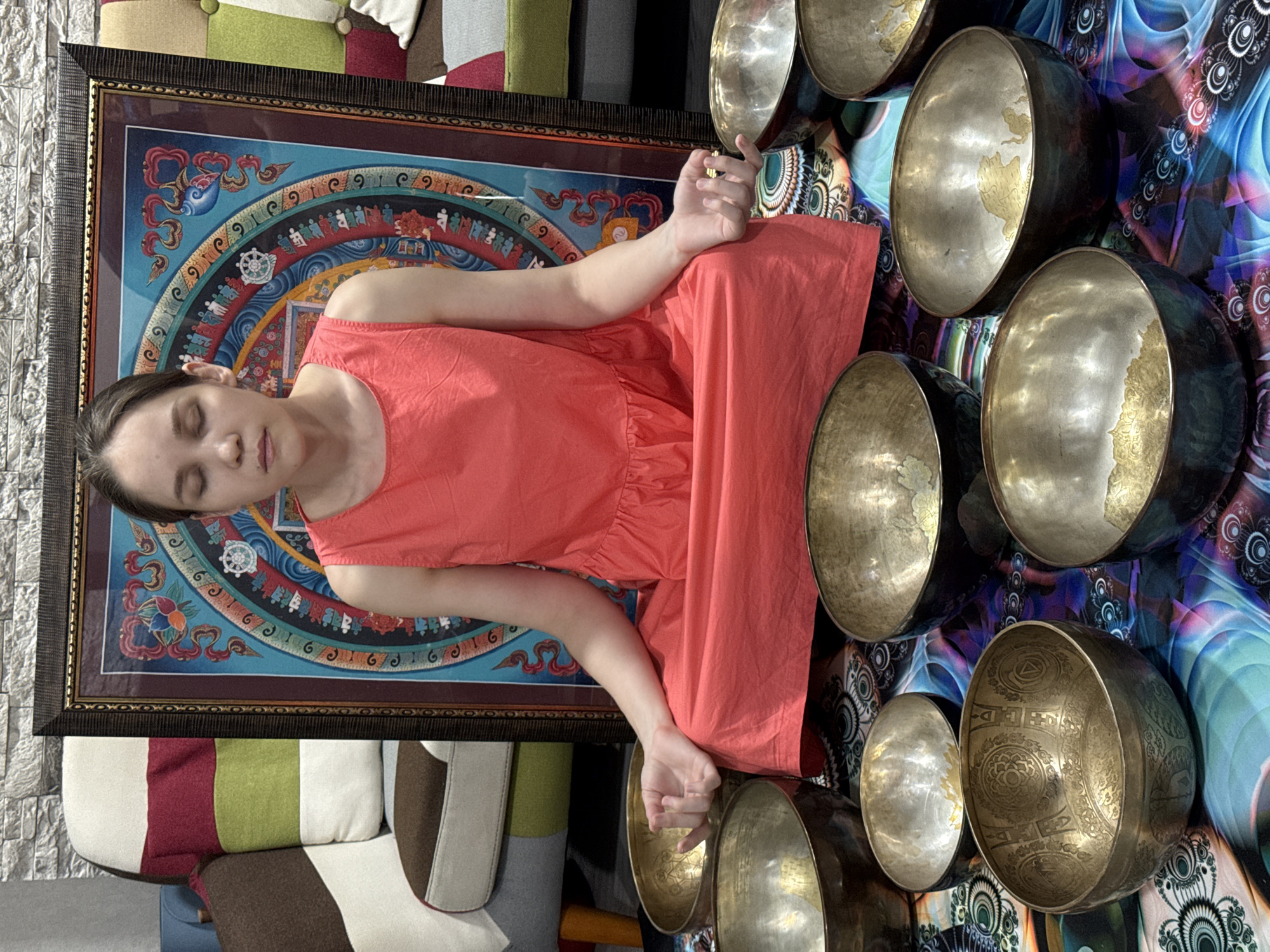 A young, light-skinned woman in a coral-colored dress sits in a meditative pose, eyes closed, on top of a framed mandala tapestry. Around her are numerous metal singing bowls of varying sizes. She appears to be indoors, possibly in a home or studio.
It is not possible to confirm the location as Tashkent, Uzbekistan from the visual data provided.