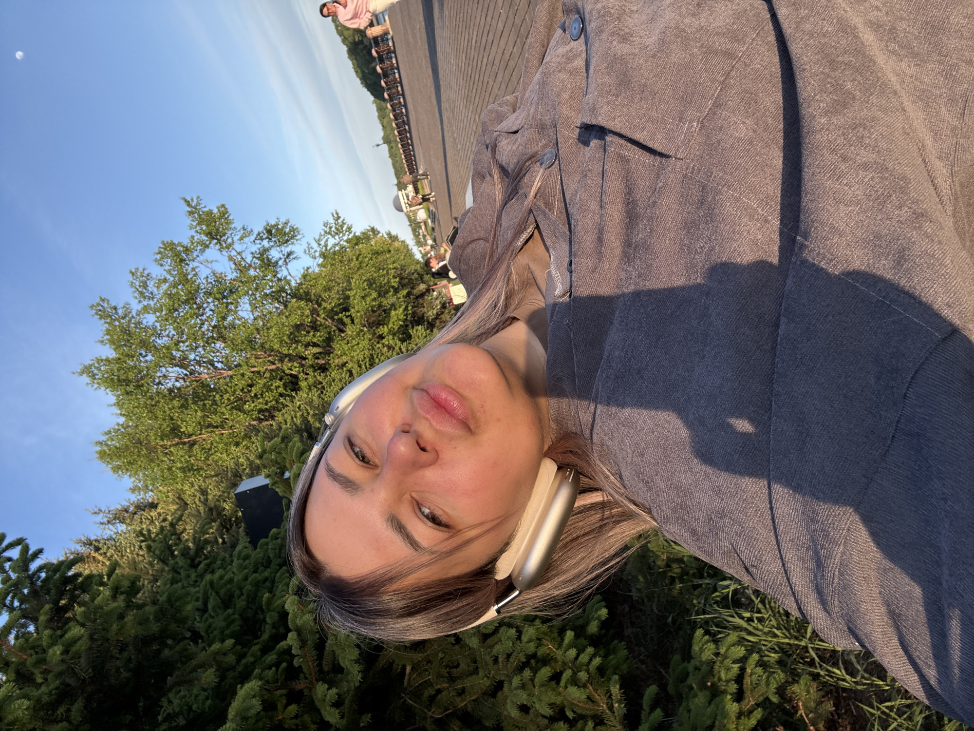 A person is wearing headphones and a gray jacket, partially framed by green vegetation. Above the trees, a blue sky is visible with a bright, circular object, likely the moon. In the background, there's a walkway with benches and people, suggesting a park or similar public space. The setting appears to be Birobidzhan, Russia.