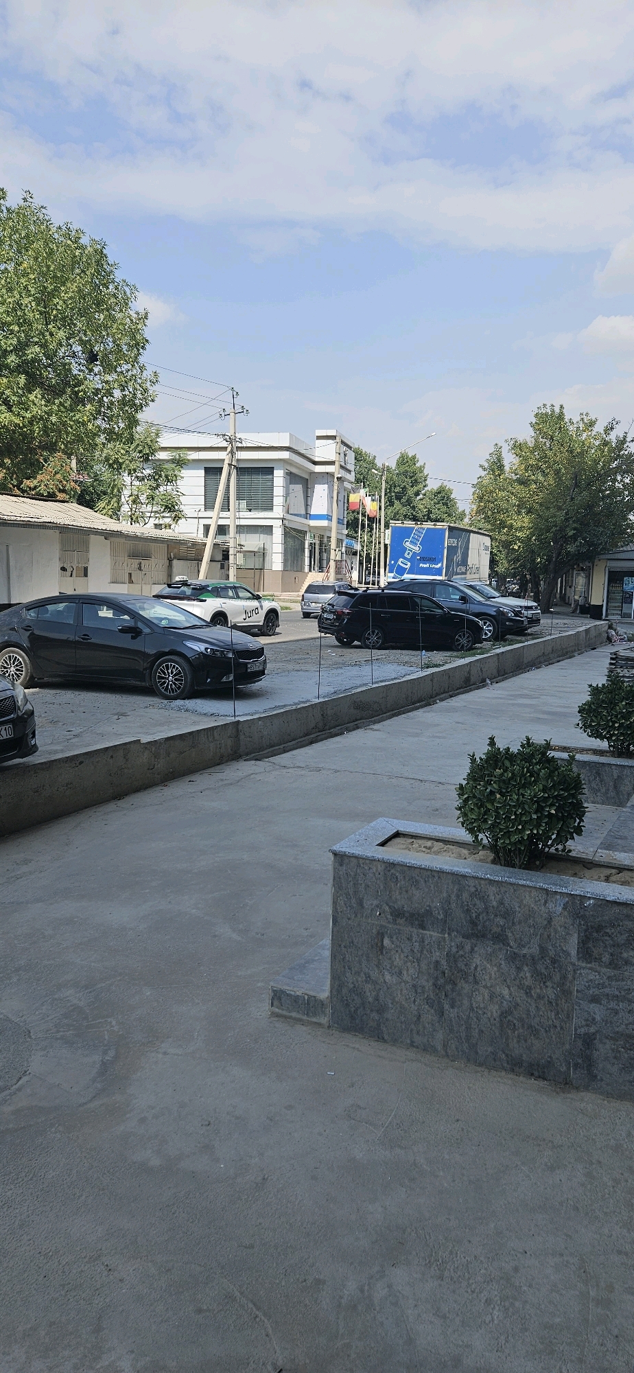 In Dushanbe, Tajikistan, a sunny day is captured in this image of a street scene. Several cars are parked along a concrete curb. A black Kia Forte is in the foreground, with its license plate clearly visible. Behind it, a white car with green accents and the word "Jura" is parked. Further down the street, a dark SUV and another black car are visible.
The background features a modern, white, multi-story building with tinted windows. Flags are mounted on a pole in front of the building, including what appear to be national flags. A large blue truck trailer with an image of a seatbelt is parked further down the street. Trees line the street, providing shade. Electrical poles and wires are also prominent features of the urban environment. The foreground shows a concrete walkway with decorative planter boxes containing small green bushes. The sky is clear with scattered clouds.