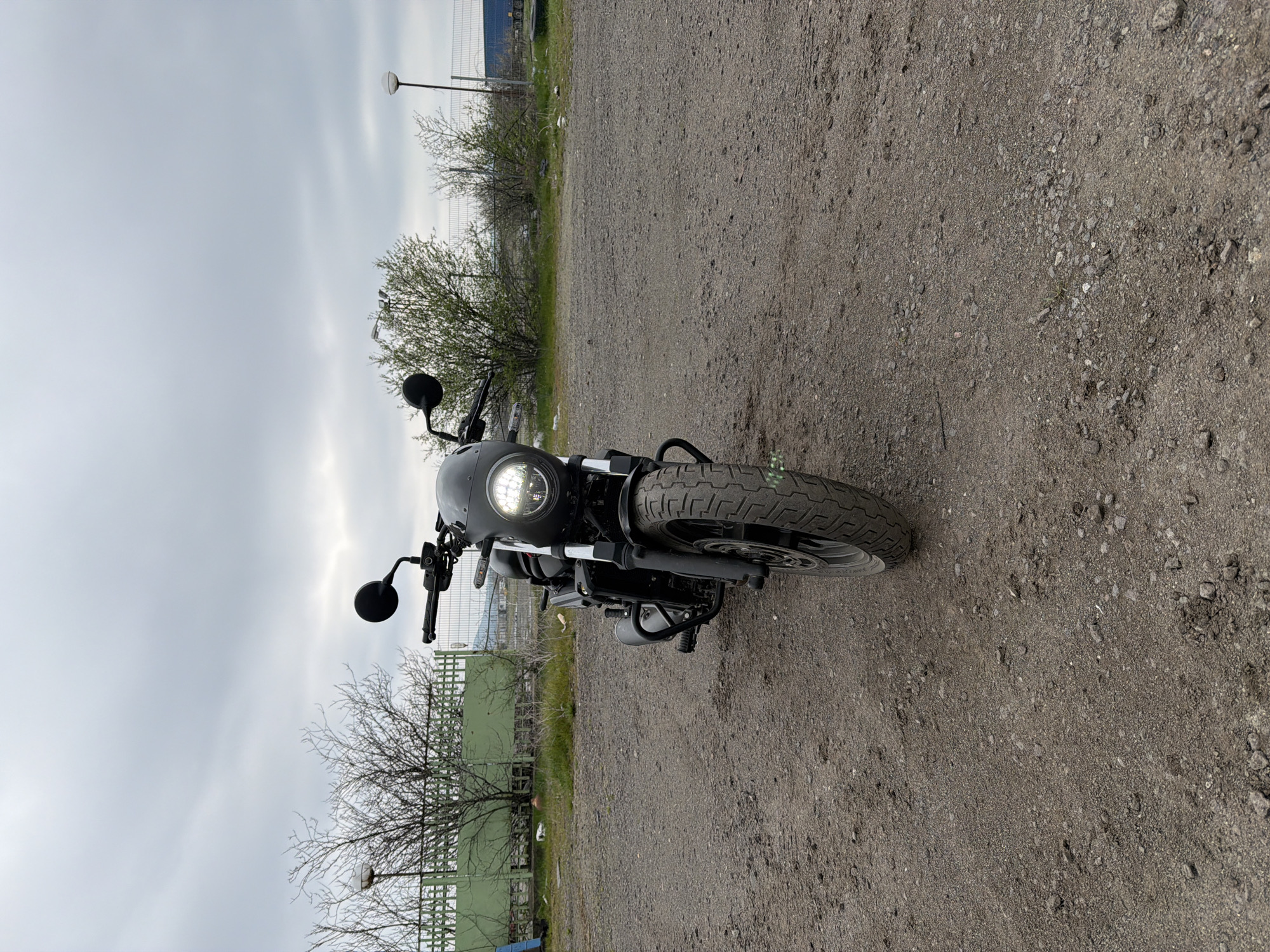 A black motorcycle is parked on a gravel lot in what appears to be an industrial area. The sky is overcast. A chain-link fence is visible in the background, as well as scrub brush and a light pole. The location is potentially in Burgas, Bulgaria.