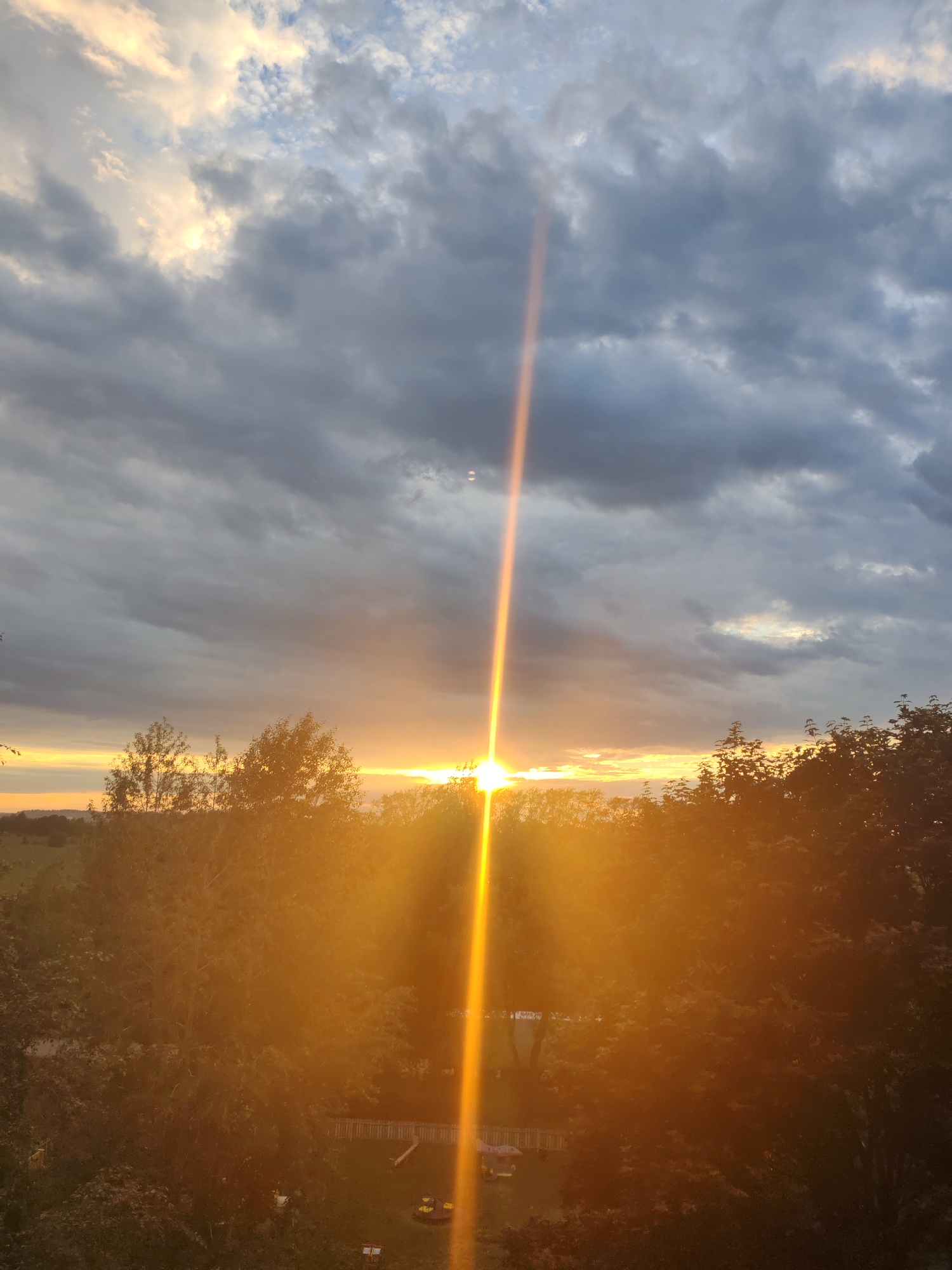The image depicts a sunset over a landscape, likely on the outskirts of Сокол, Russia. The sky is filled with a dramatic mix of dark, heavy clouds and patches of lighter, illuminated sky where the sun is breaking through. A prominent beam of golden sunlight shoots vertically upwards from the horizon, bisecting the scene. Below the sky, a dense line of trees forms a silhouette against the vibrant sunset. The trees are predominantly green and brown, with the sunlight creating a strong lens flare effect that obscures some of the details. In the lower portion of the image, hints of a residential area are visible, including a fenced yard with some playground equipment, suggesting a suburban or semi-rural setting. The overall impression is one of natural beauty and a tranquil evening setting.